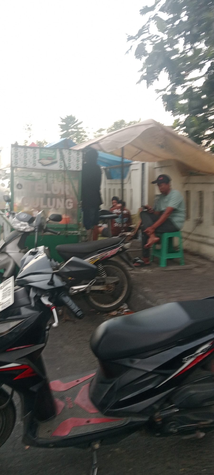 The image depicts an outdoor scene, likely a street-side vendor stall, with several motorcycles parked nearby. In the foreground, the rear section of a black and red scooter is visible. Behind it, a black and silver motorcycle is parked. Further back, a food stall is set up with a light-colored awning. The stall has green paneling with white text and graphics. A person wearing a dark shirt and dark pants stands partially obscured in front of the stall. To the right of the stall, a man wearing a light green t-shirt, dark pants, and a black cap sits on a green stool, holding a mobile phone. Two other individuals are seated behind a table near the stall. The environment appears to be urban, with trees visible in the background and a hazy sky, suggesting it may be early morning or late afternoon. The scene is consistent with a typical street food vendor location in a city like Ngemplak, Indonesia, where motorcycles are a common mode of transportation.