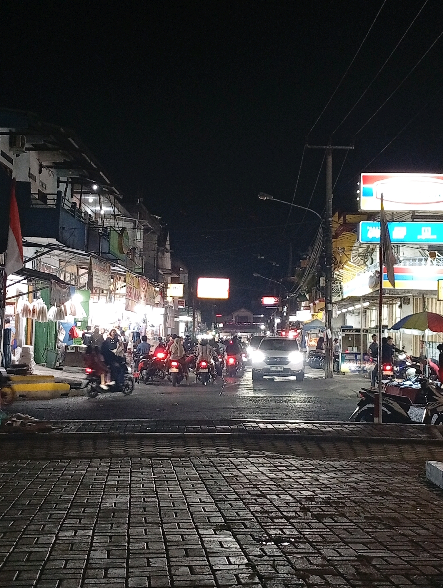 The image depicts a bustling street scene at night in Cileungsi, Indonesia. The foreground is dominated by a textured, brick-paved walkway with puddles reflecting the ambient light. Beyond the walkway, a street is filled with activity. Numerous motorcycles, many with illuminated taillights, are in motion, suggesting traffic. One white car with its headlights on is visible in the middle of the street. On either side of the street are buildings with storefronts illuminated by bright lights. Signs are visible on the buildings, some featuring text and logos, indicating commercial establishments. Several people are seen on motorcycles and walking near the storefronts. Overhead, power lines and a street lamp are present. The overall atmosphere is one of a lively, urban night market or commercial district.