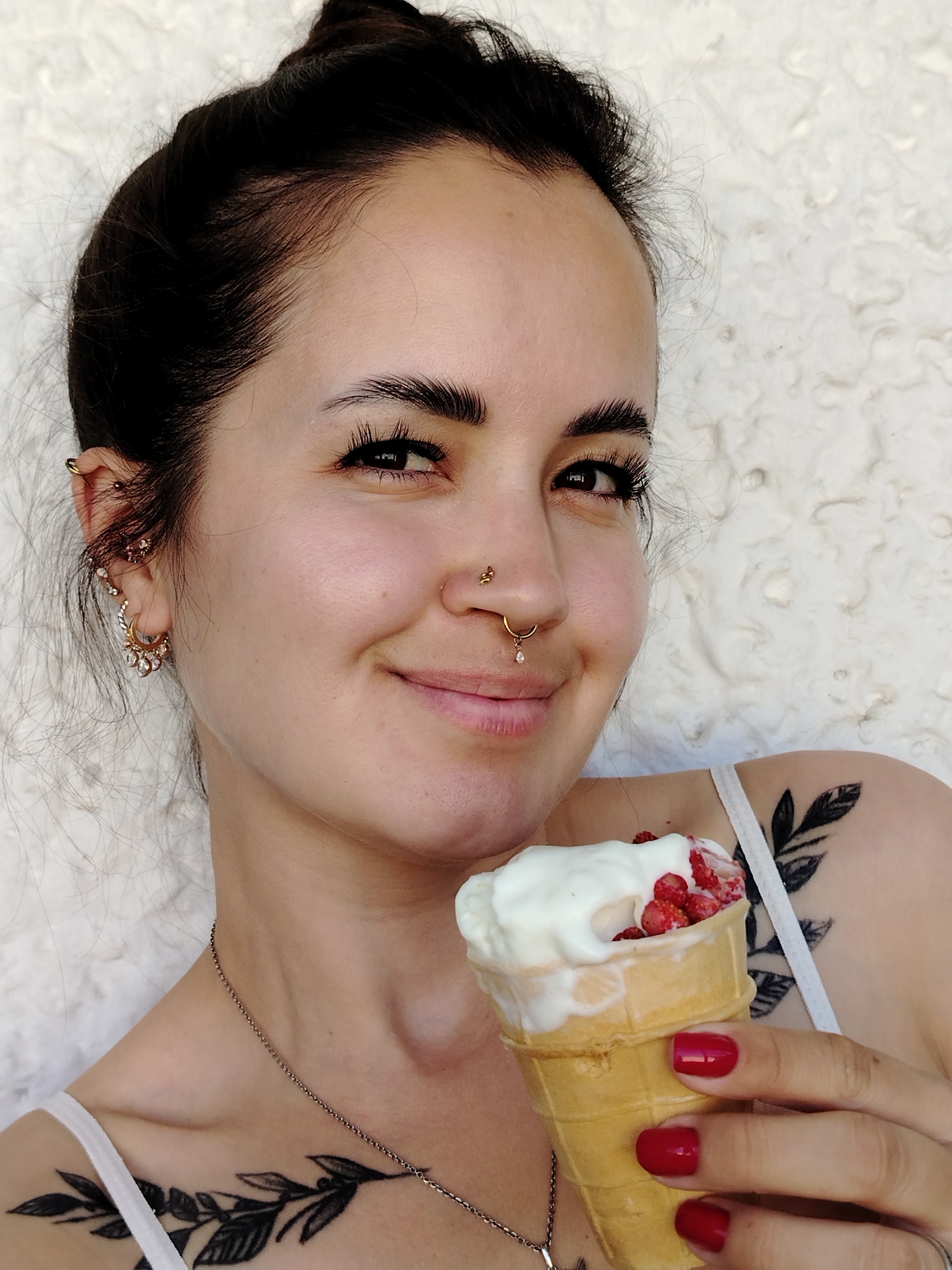 A close-up medium shot shows a woman holding an ice cream cone and smiling at the camera. She has dark hair pulled back in a bun, dark eyebrows, and is wearing several pieces of jewelry, including hoop earrings, a nose ring, and a lip piercing. She is also wearing a white tank top, and has tattoos on her shoulder and chest. The ice cream cone is vanilla flavored with strawberries. The background is a textured white wall. The lighting suggests it is daytime.