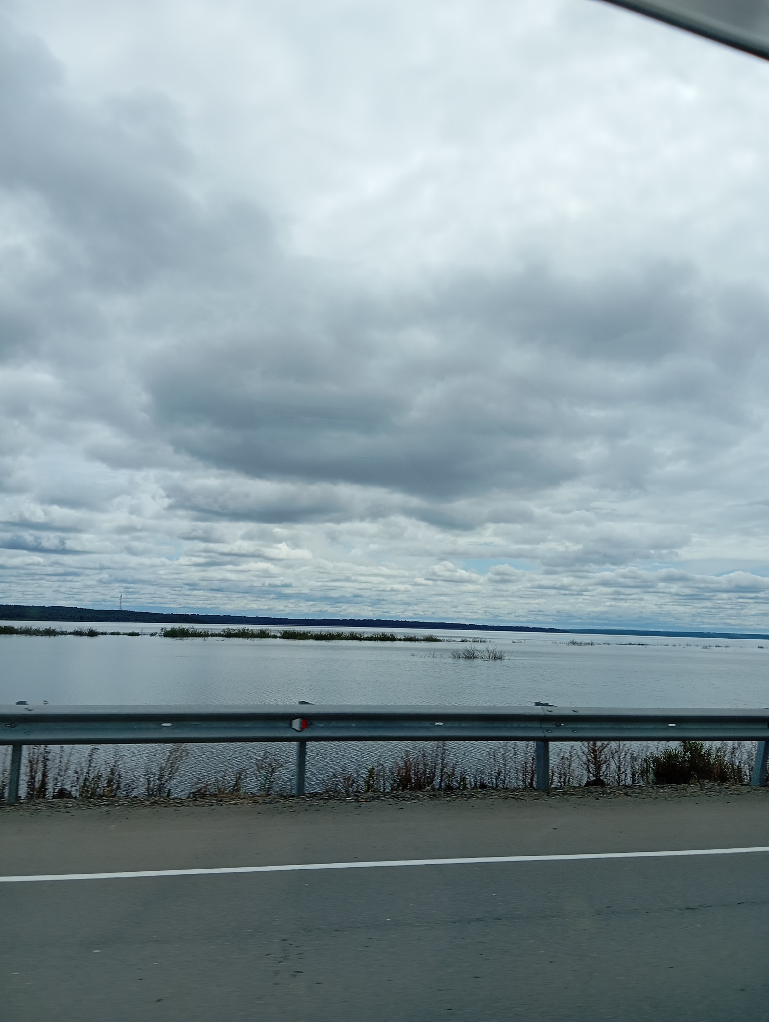 This is a vertical, eye-level, medium shot of a cloudy sky over a body of water. The sky is dominated by a dense layer of gray clouds, with some lighter patches visible in the distance. A calm body of water stretches across the middle ground, reflecting the muted tones of the sky. In the far distance, a thin, dark line of trees or land marks the horizon. In the foreground, a section of asphalt road is visible, with a white line marking the lane. A metal guardrail runs along the edge of the road, separating it from the embankment of dry grass and thin trees. The scene appears to be a roadside view, likely from a vehicle, looking out towards a natural landscape. The location is the city of Mayak, Russia.