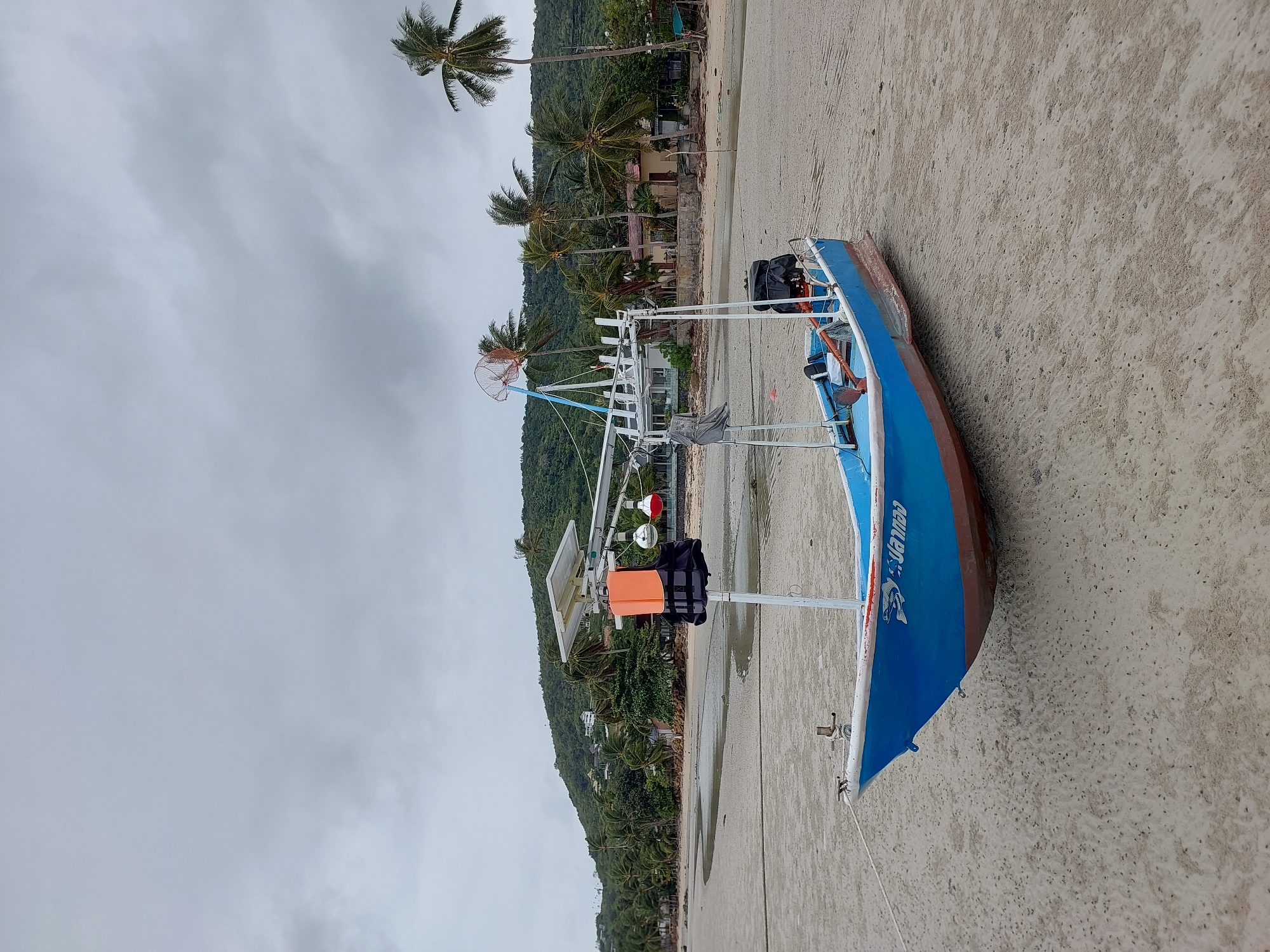 A blue and red fishing boat rests on a sandy beach under a cloudy sky. The boat has a white frame structure above it, with a solar panel, a life jacket, and two red fishing floats attached. A life vest is hanging on the structure. The beach is wet and appears to be at low tide, with small channels of water visible. In the background, a lush green hill is covered in palm trees and scattered buildings. The scene is located on the island of Ko Samui in Thailand.