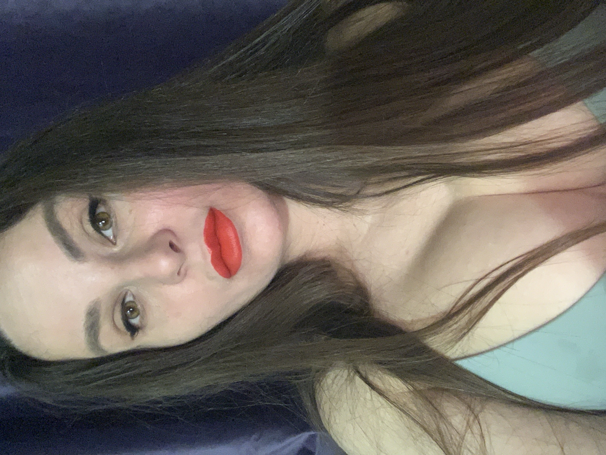 A woman with long, dark brown hair lies against a dark blue or purple surface, likely fabric. Her face is angled towards the camera, revealing fair skin, green eyes, and red lipstick. Her clothing appears to be light blue or green. The location is unknown.