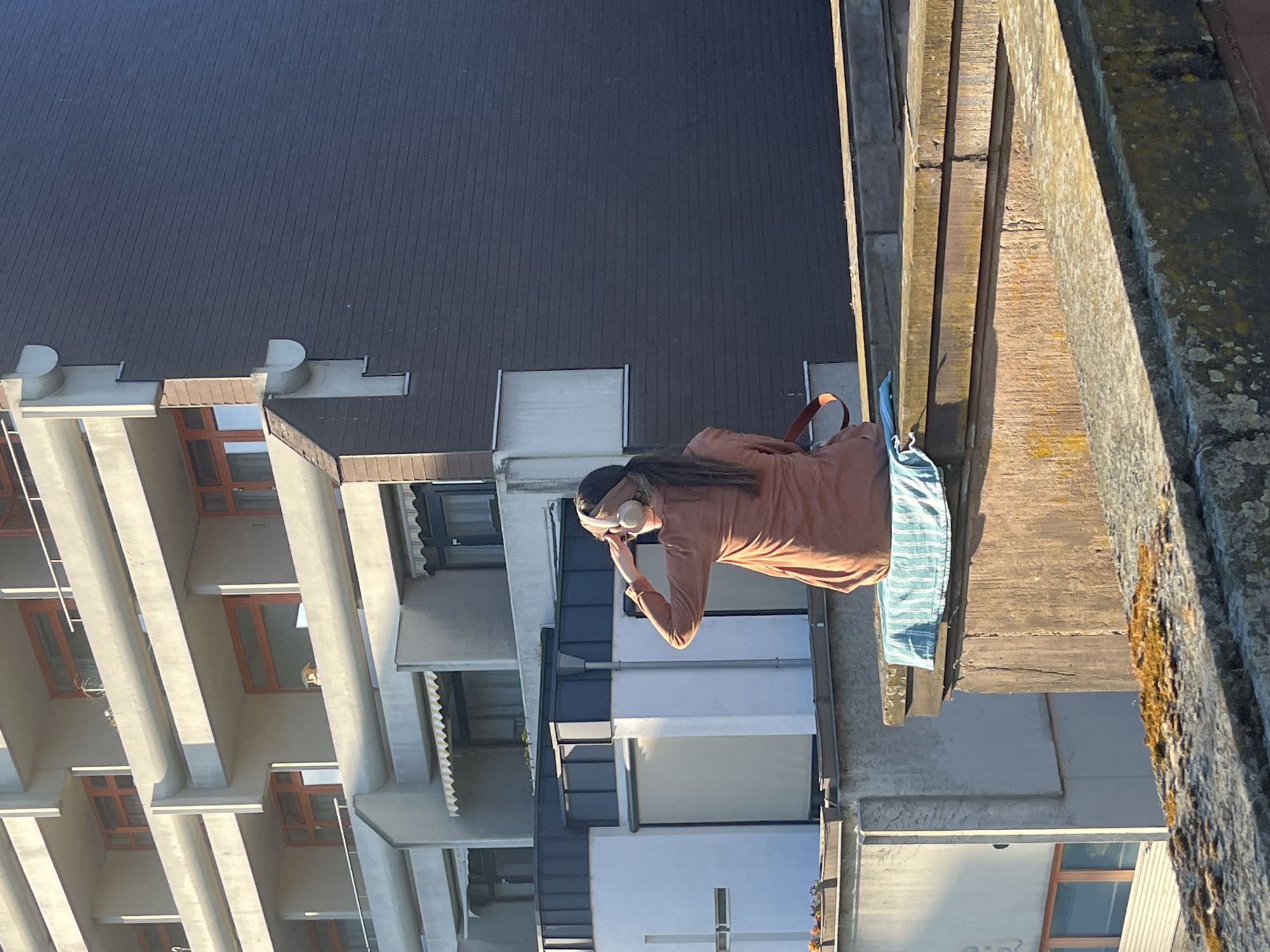 A medium-angle, outdoor shot captures a person wearing an earth-tone shirt and headphones standing on a balcony, with a blue bag nearby. The background reveals a modern building with protruding balconies and orange-trimmed windows. To the right of the balcony, a moss-covered wall is visible. The photograph appears to have been taken in Saarbrücken, Germany.