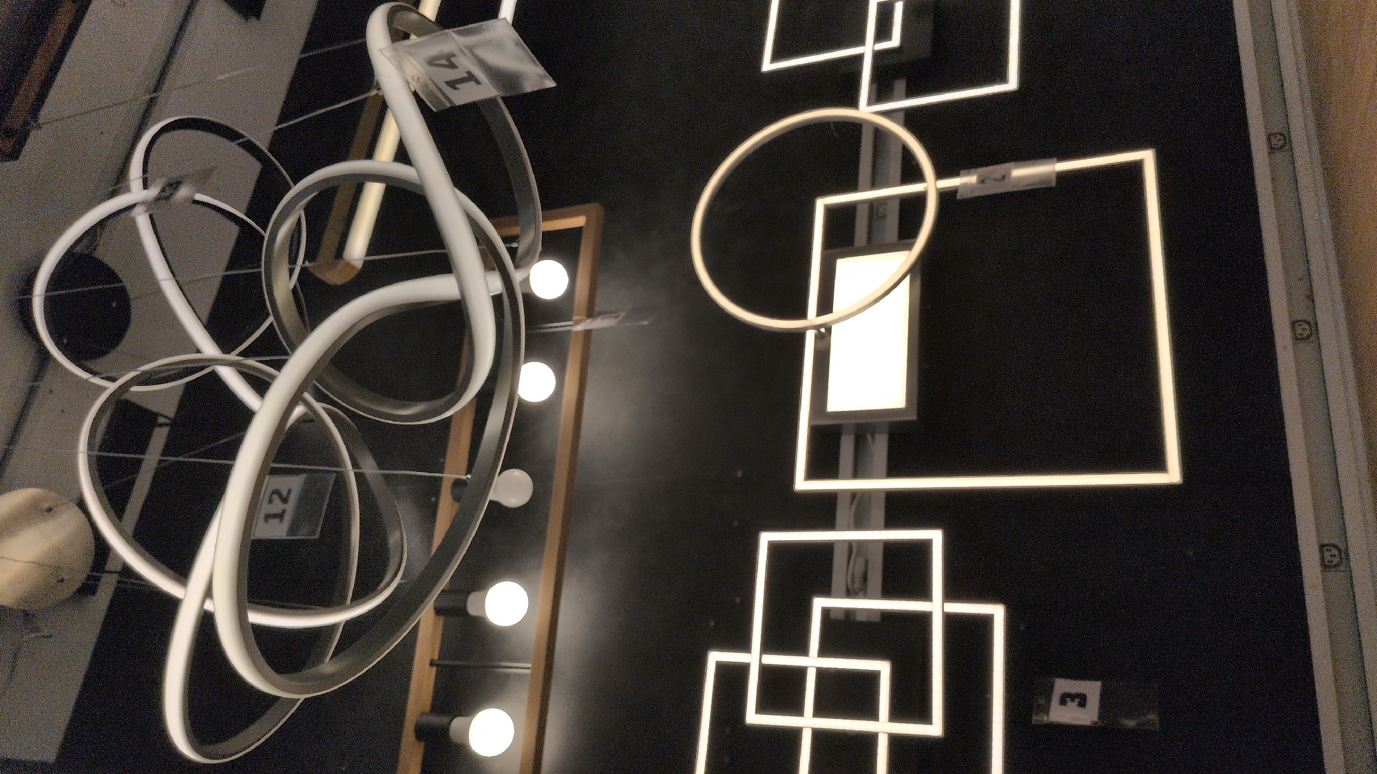 The image displays a retail environment, likely a lighting store, with several modern, illuminated fixtures. In the upper section, a large, multi-ringed pendant light with white LED elements is prominently featured, with price tags labeled "12" and "14" attached to it. Below this, a row of five exposed light bulbs hang from the ceiling, illuminating a wooden plank.
The lower section of the image showcases geometric LED lighting designs mounted on a dark wall. These include a series of interconnected squares and a circular ring light, all emitting a bright white glow. A price tag labeled "3" is visible near the bottom left, attached to a square fixture. Another tag with the number "2" is on the circular light. The overall impression is of a well-lit showroom displaying contemporary lighting solutions. There are no people visible in the image. The environment appears to be an indoor commercial space, with a hint of shelving or display units at the very bottom. There is no information in the image to suggest a specific location in the city of Sömmerda, Germany.
