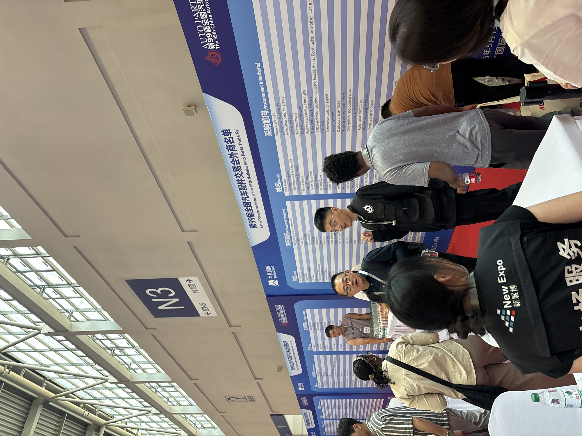 The image shows a crowd of people at the 99th China Auto Parts Trade Fair, held in an event space marked as "N3" within the Liangjiang area of an unspecified city in China. Attendees are examining exhibitor booths, specifically lists of foreign investors and their procurement intentions. The lists include auto parts such as wheels, tires, headlights, bumpers, engines, rubber products, and accessories. Some individuals appear to be employees, identifiable by "New Expo" vests. The scene is indoors with a visible ceiling structure.