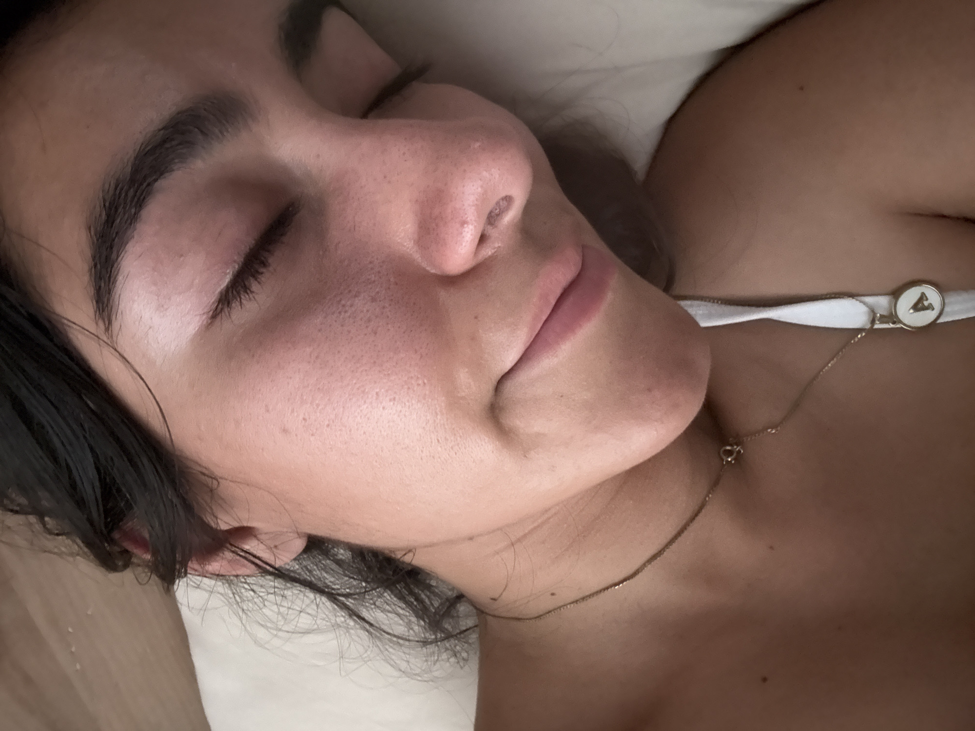 The image captures a close-up, slightly angled view of a young woman resting, possibly asleep or deep in thought. She is positioned on her right side, with her head resting on a light-colored, soft surface, likely a pillow. Her eyes are closed, and her facial expression appears serene and peaceful.
Her dark hair frames the side of her face, with some strands falling near her ear and neck. Her skin has a warm, natural tone, showcasing visible pores, particularly on her nose and cheek, suggesting a natural and unedited appearance. She has well-defined, dark eyebrows and long, dark eyelashes. Her lips are slightly parted, revealing their natural pink hue.
She is wearing a delicate, gold-colored chain necklace with a small heart-shaped clasp visible near her neck. Further down, near her left shoulder, another part of the necklace drapes over a white strap of clothing, which appears to be part of a top or brassiere. Attached to this white strap is a small, round pendant with a white background and a distinct dark symbol, possibly a stylized letter "A" or a triangular shape, similar to the Vivienne Westwood orb logo. The lighting is soft and even, suggesting an indoor setting, without strong shadows or bright natural light, making the exact time of day indeterminate. The focus is sharp on her face and upper body.