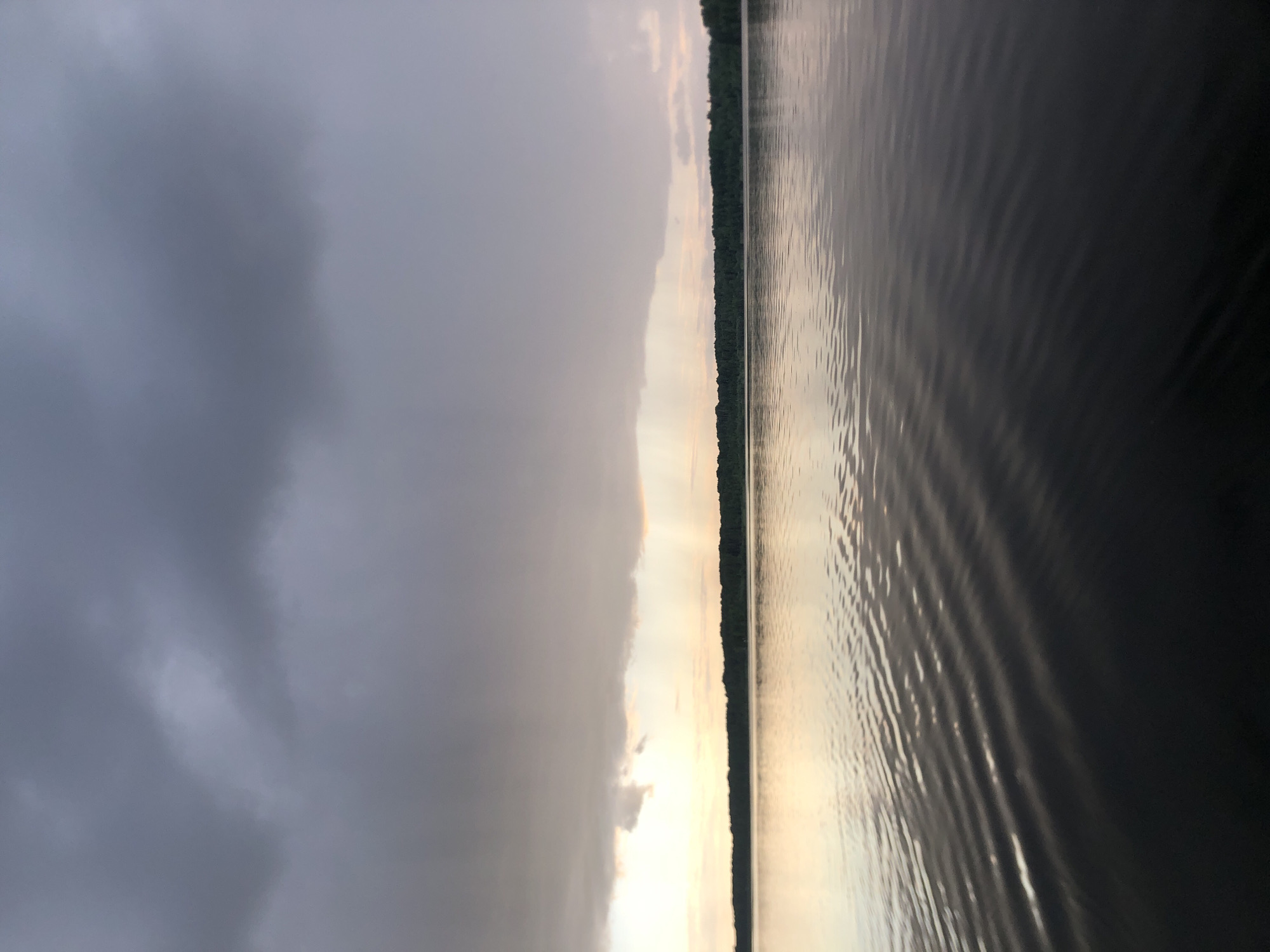 This is a landscape photograph taken from a watercraft on a lake or large body of water, likely near Svirstroy, Russia. The image is oriented vertically. The scene is divided into two distinct halves: the left side is dominated by dark, heavy clouds, suggesting an overcast or approaching storm. The right side of the image features a calm body of water with visible ripples reflecting the soft, warm light of either sunrise or sunset. A thin, dark line of trees along the distant shore separates the sky from the water. The lighting indicates it is either early morning or late evening. There is no visible text in the image. The overall mood is serene and perhaps a little dramatic due to the contrast between the stormy sky and the peaceful water.