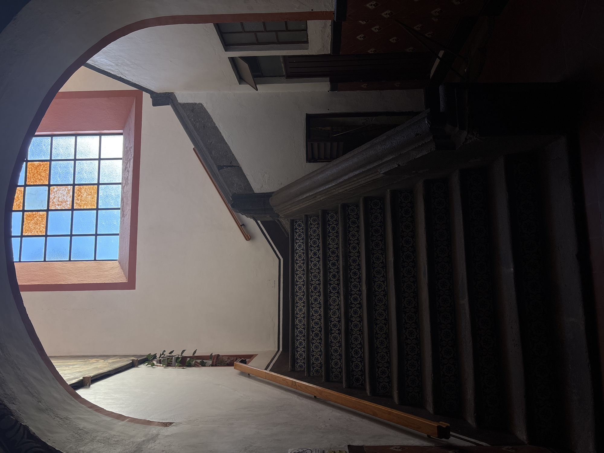 The image depicts the interior of a building in Puebla, Mexico, looking up a staircase. The staircase has patterned blue and white tiles on the risers. A handrail runs along the bottom of the staircase. A window with a gridded design and panes of alternating clear and orange tinted glass is visible to the left. The walls are white and the lighting is dim, creating shadows on the stairs and walls. The scene is viewed through an archway.