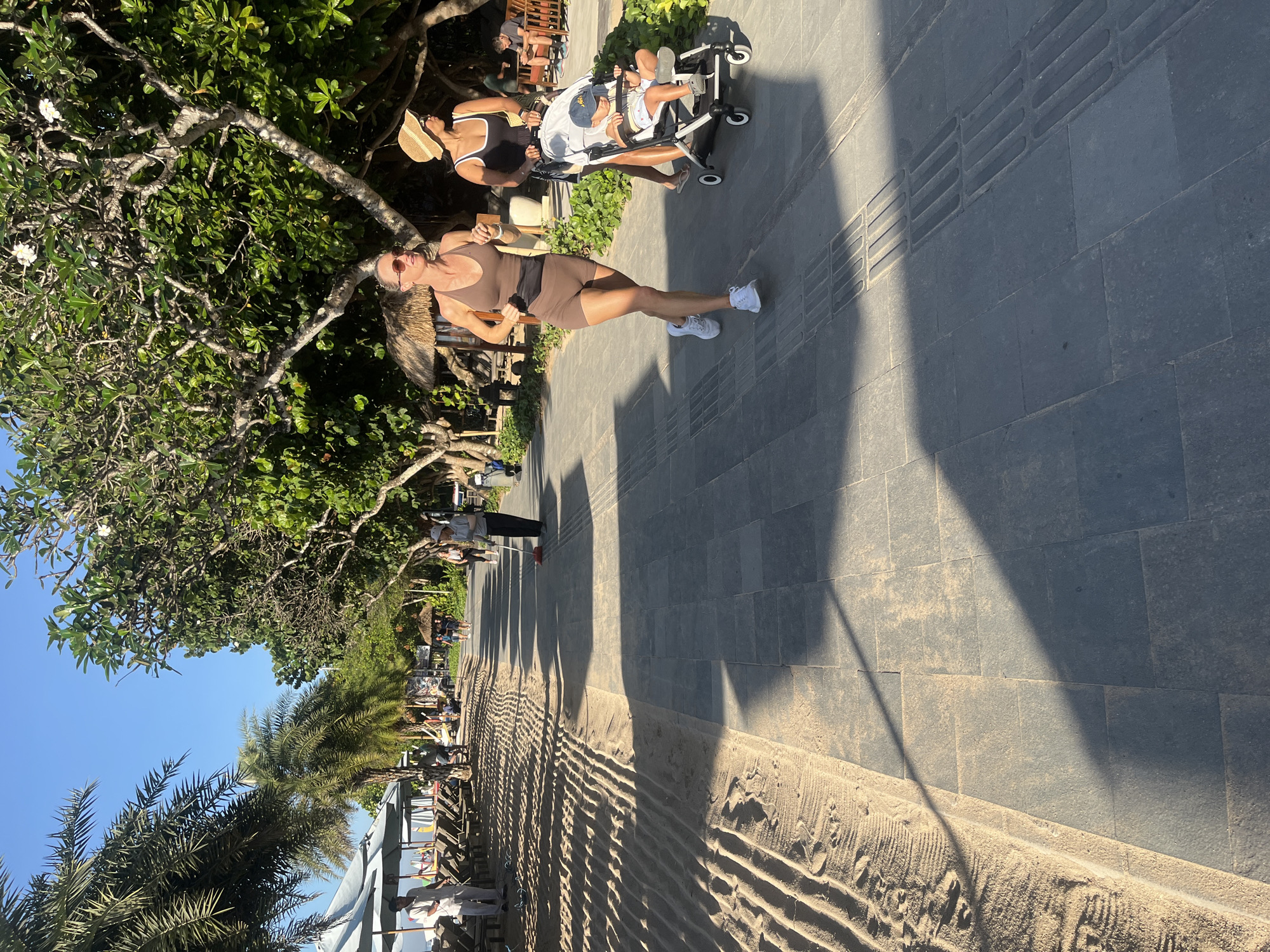 Two women are walking on a paved path beside a sandy area lined with palm trees. The woman on the left, wearing a brown athletic outfit, is jogging. The woman on the right, wearing a black tank top and straw hat, is pushing a stroller with a baby inside. The background features more palm trees, some lounge chairs, and umbrellas, suggesting a beach resort or public beach area in Denpasar, Indonesia. The sun is shining, casting long shadows on the path. A person is seen sweeping the path in the distance.