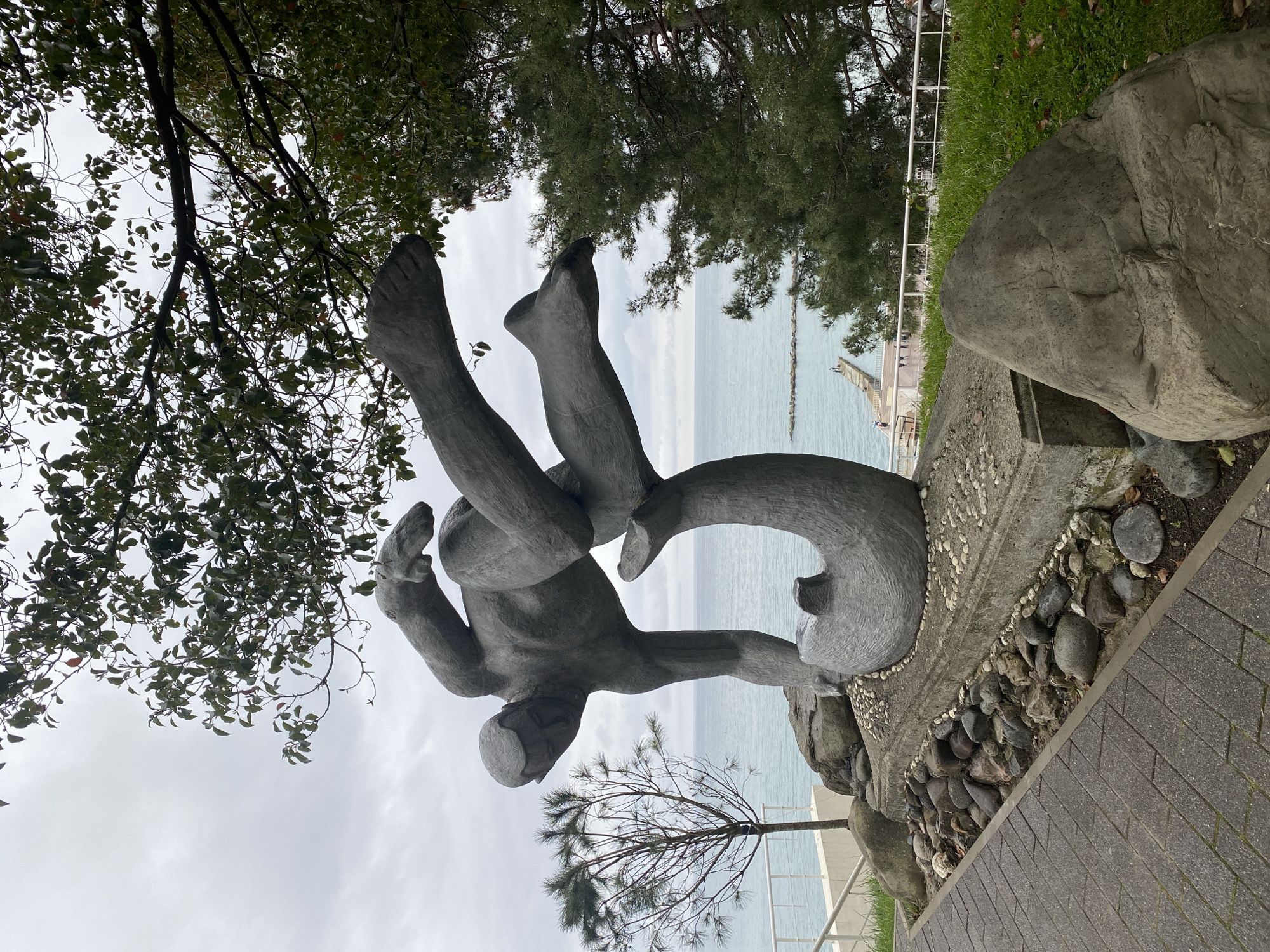 The image captures a sculptural composition located near the Black Sea in Sochi, Russia. The statue, rendered in gray, depicts a stylized human figure holding a pair of abstract forms resembling dolphins. The sculpture rests on a rocky base adorned with small stones and is bordered by a paved walkway. In the background, the sea stretches to the horizon, with a pier visible in the distance. Trees and foliage frame the scene on both sides and overhead. The sky appears overcast.