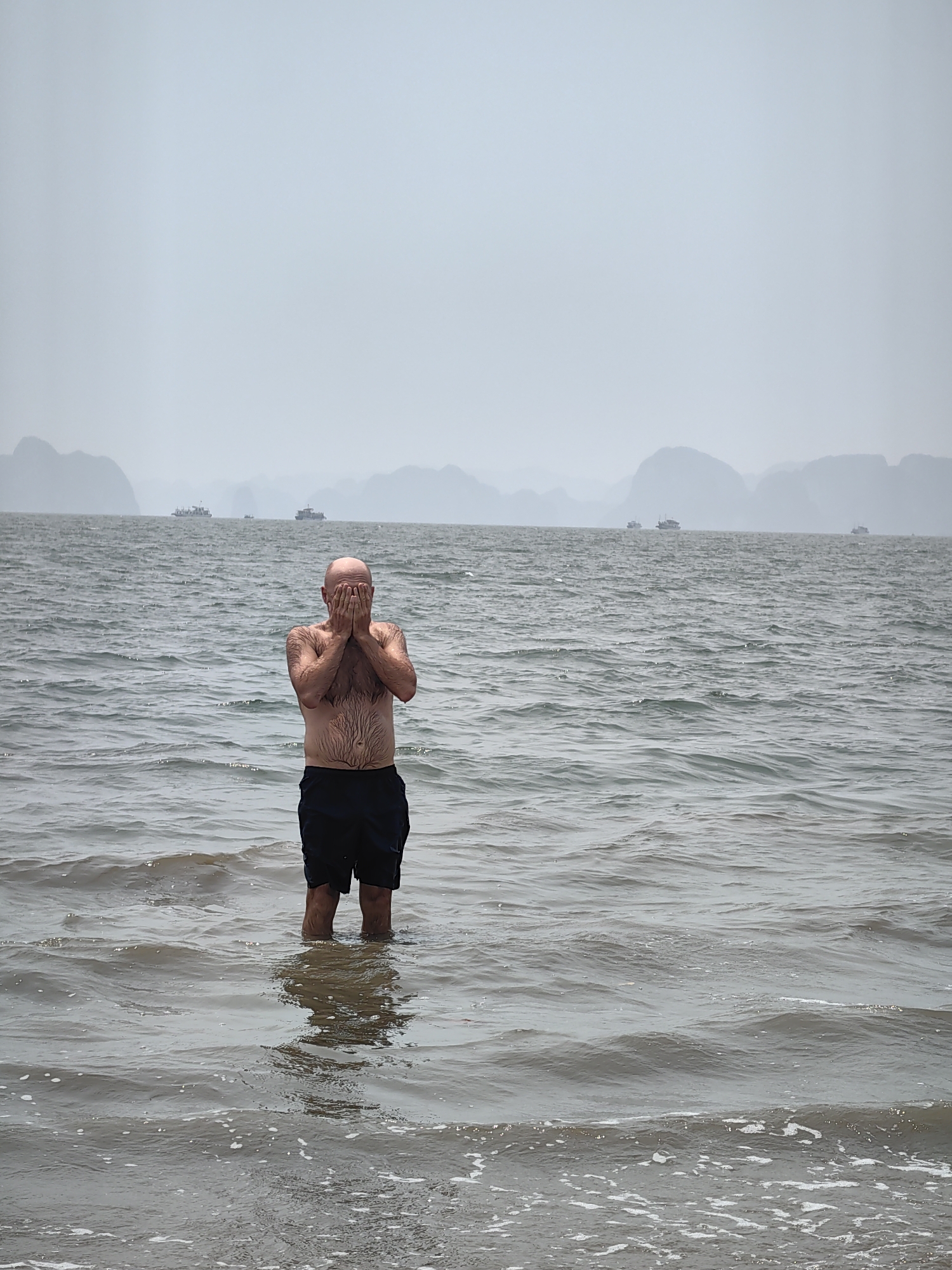 A bald man in black shorts stands waist-deep in the water of Bai Chay Ward, Vietnam. He is facing the camera with both hands covering his face. In the background, a grey, hazy seascape shows several boats on the water and rocky islands in the distance. The water is choppy with small waves.