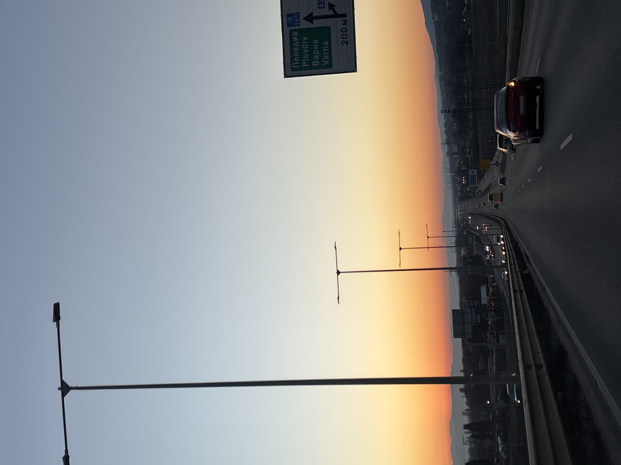 The image shows a highway during sunset. The highway sign indicates directions to Plovdiv and Varna, 200 meters away. Cars are driving on the highway, and streetlights line the road. The sky transitions from blue to orange. The location appears to be near Sofia, Bulgaria, based on the highway sign destinations. A red car is visible, near the camera.