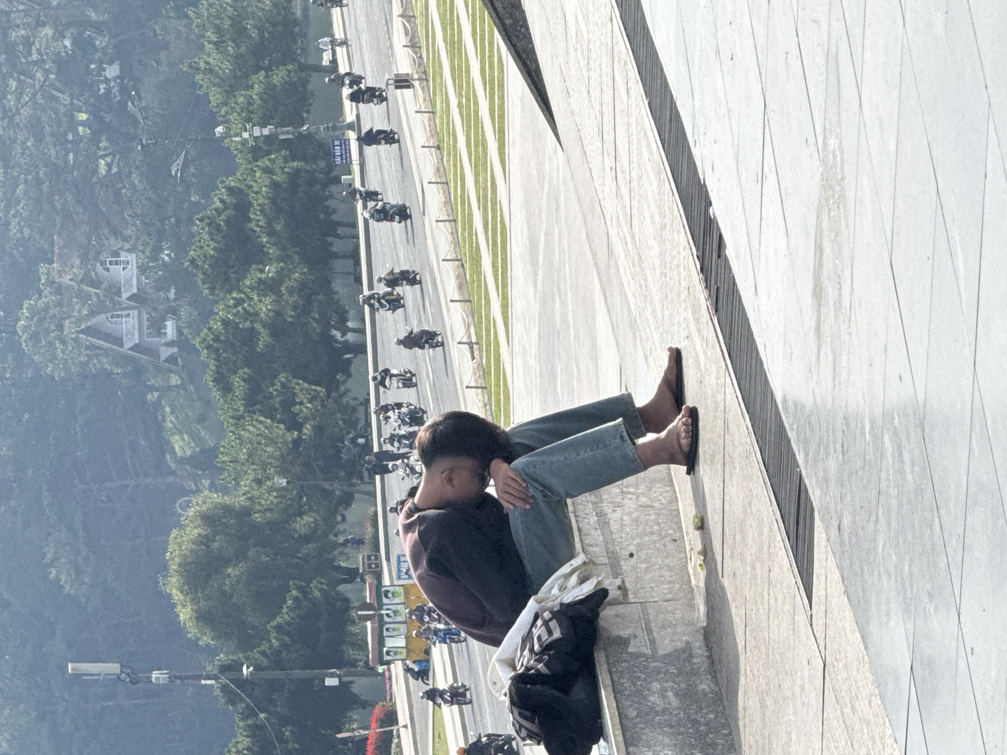 A person sits on the edge of a large concrete structure, positioned with their back towards a street in Da Lat, Vietnam, marked by heavy motorbike traffic. The individual is wearing jeans, a dark-colored sweatshirt, and flip-flops. A bag rests near them on the ledge. The backdrop includes trees and houses on a hillside, along with lampposts lining the road.