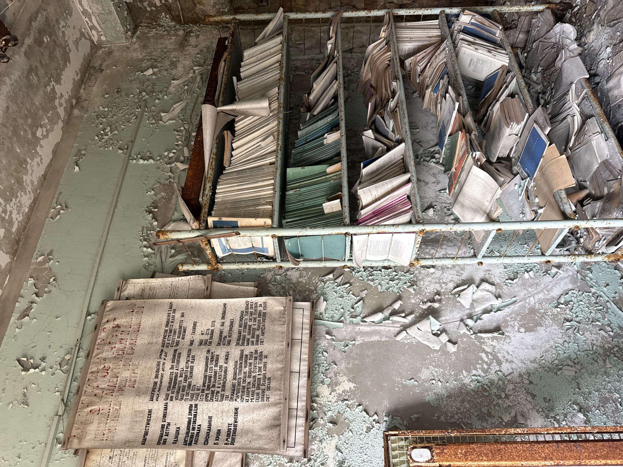 This indoor shot depicts a derelict room, likely in Prypiat, Ukraine, showing significant deterioration. Flaking paint peels from the walls and floor. The focus is on a rusty metal shelving unit filled with stacks of disorganized documents, files, and books. Several large, faded, and text-heavy sheets are positioned on the floor in front of the shelving unit. A rusty heater element is partially visible in the lower right corner. The overall environment suggests abandonment and neglect.