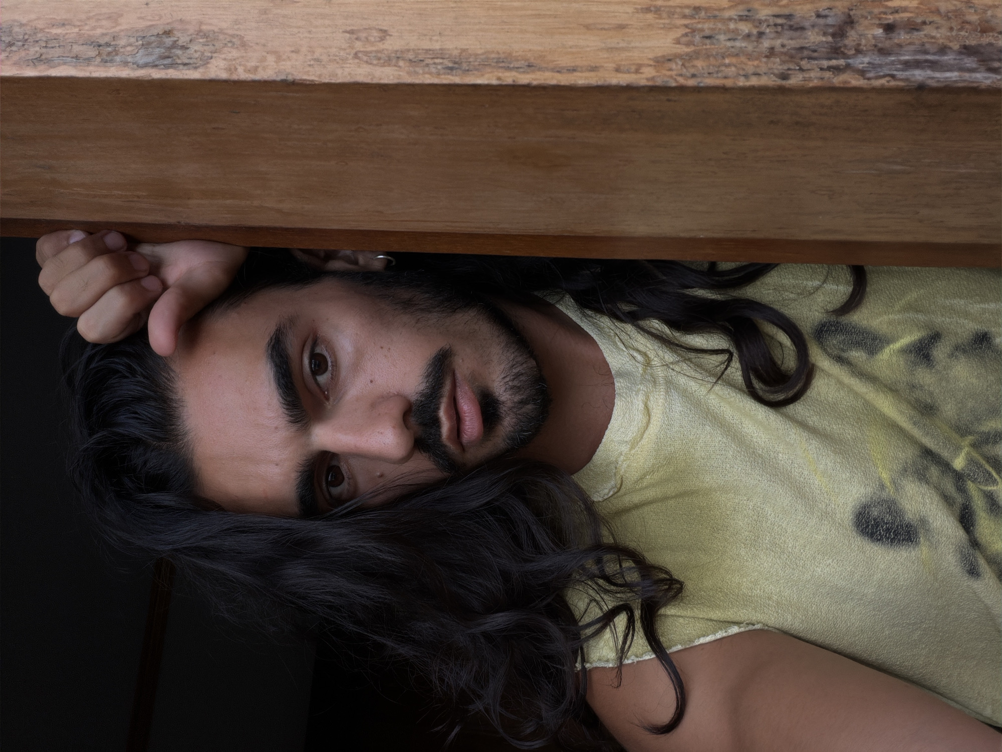 The image is a close-up, intimate portrait of a person, likely male, positioned sideways in the frame. The subject has long, dark, wavy hair that frames their face, along with a full beard and mustache. They possess dark, piercing eyes and prominent eyebrows, maintaining direct eye contact with the camera.
The person is wearing a light yellow or chartreuse-colored t-shirt, which features subtle, faded dark patterns or designs. A sturdy wooden surface, possibly a shelf or beam, occupies the top portion of the image, running horizontally above the subject's head. One of the person's hands is visible, resting on this wooden edge near their temple. The background to the subject's left is dark, indicating a shaded area or low light, contrasting with the softly lit face and clothing. The pose appears relaxed or contemplative.
No specific activity beyond posing for the portrait is evident. The overall impression is one of calm and direct engagement. The setting is implied to be indoors, and the provided location context is North Kuta, Indonesia. There is no visible text within the image.