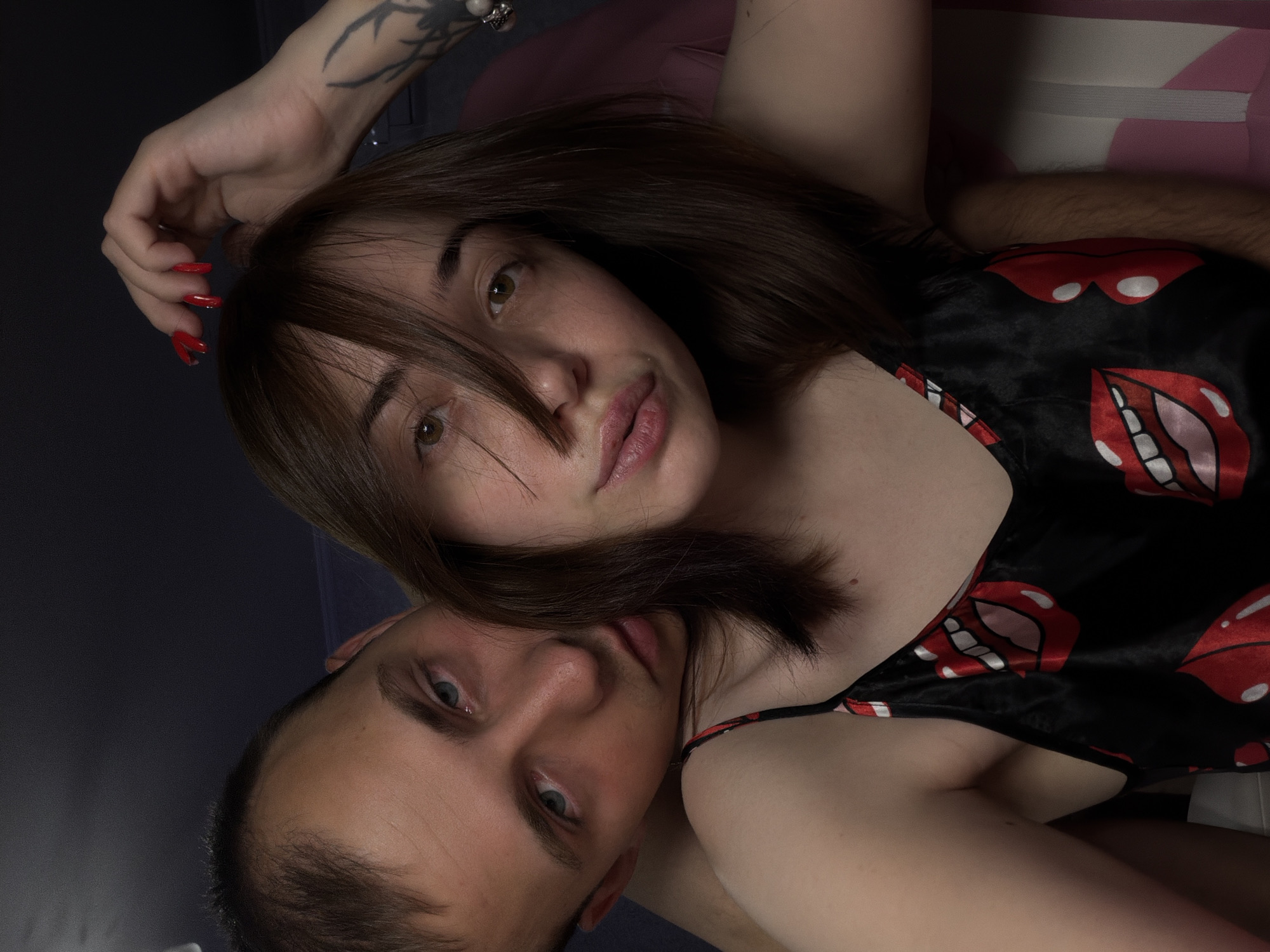 This media file shows a close-up, overhead shot of two people, a man and a woman, lying down. The image is rotated, with the man's face positioned in the lower left and the woman's face in the upper center.
The woman has brown hair that falls over her face and onto her chest. She is looking towards the camera with her eyes slightly open. Her lips are parted, and she is wearing a black garment with a pattern of red lips. Her left hand, with long red nails, is raised and resting near her head, which has a tattoo on it.
The man's face is visible in the lower part of the frame. He has light blue eyes and short brown hair. His head is partially obscured by the woman's hair and body. He is also looking towards the camera.
The background is dark and appears to be indoors. There is a hint of a patterned surface, possibly a pillow or bedding, behind the woman's head, with white and pink stripes. The lighting is soft, suggesting it might be indoors at an unspecified time of day. The overall mood appears intimate and relaxed. No text is visible in the image.