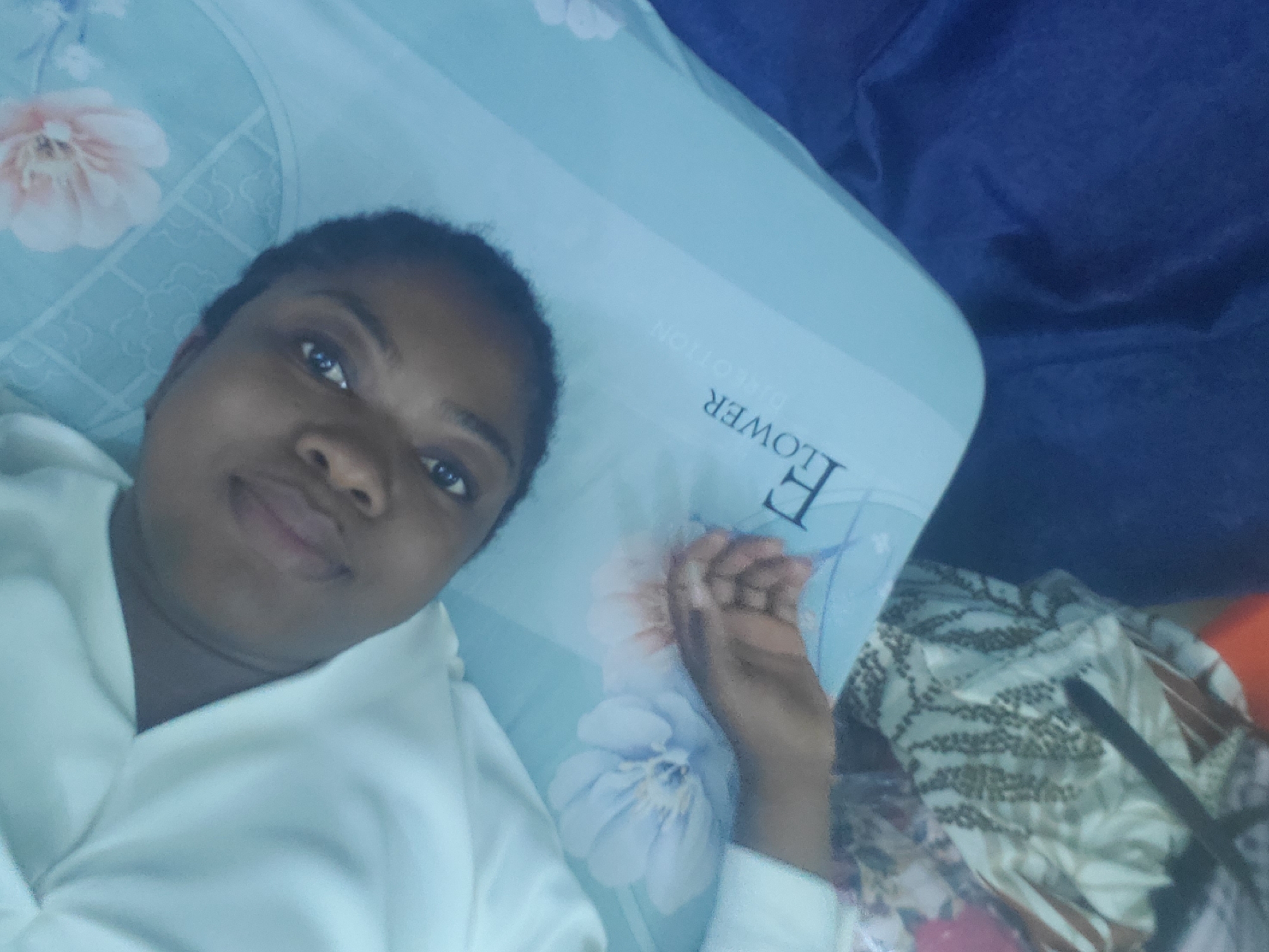 A young woman with dark skin and short dark hair lies on her back, looking directly at the camera with a gentle, relaxed smile. She is wearing a light-colored, possibly white or off-white, casual top with a hood or collar, and her right hand is visible near her head, fingers slightly curled.
The setting appears to be indoors, likely a bedroom in Barden Yana, Nigeria. She is resting on a light blue pillow or sheet adorned with delicate floral patterns in pink and light blue. Visible on the pillow, rotated ninety degrees clockwise, are the words "F FLOWER DIRECTION". To her right, a section of dark blue fabric, possibly a blanket or part of the bed, is visible. Further to the right and slightly below, there's another patterned fabric in shades of brown, white, and black, alongside a dark, slender object, possibly a pen or remote. The lighting is soft, suggesting an indoor environment, perhaps during the day or evening. The overall mood conveyed by her expression is one of calm and contentment, consistent with someone relaxing at home. The activity appears to be a self-portrait or selfie.