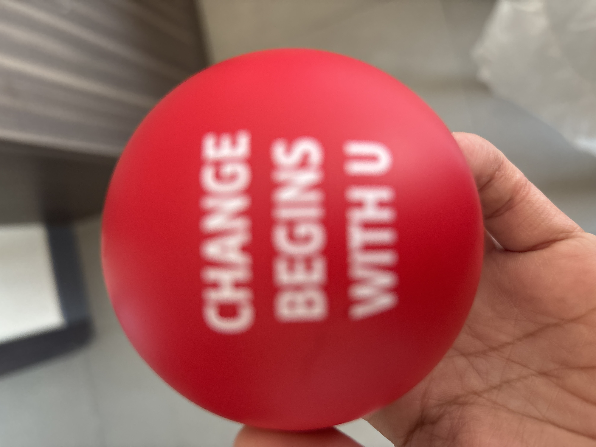 A red stress ball with white text reads "CHANGE BEGINS WITH U" is held in a person's hand. The background is blurred and shows a tiled floor and a wall. The image does not contain any information about the city of Dubai, United Arab Emirates.
