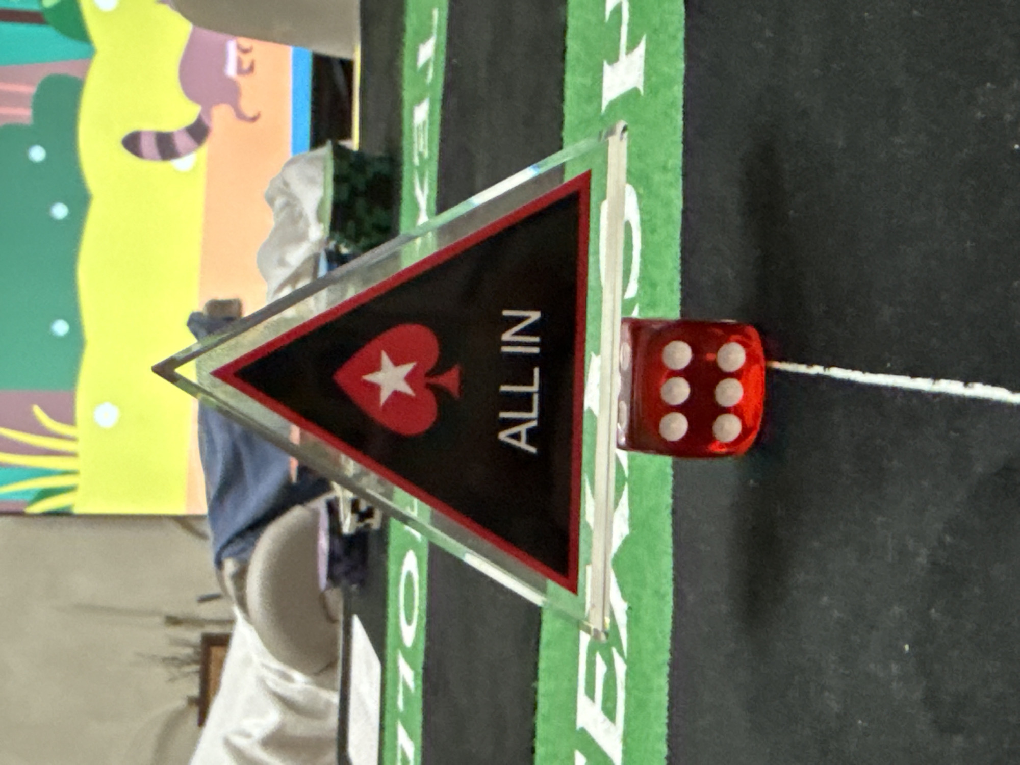 This scene, located in the city of Krasnoyarsk, Russia, presents a detailed view of a poker game setup. In the center foreground, a clear acrylic triangular poker marker is positioned, displaying a red-and-white spade symbol on a black background with the text "ALL IN" in white capital letters. Directly in front of the marker, a red six-sided die with white pips shows a '6' (three pips on its top face and three on its visible side). These objects rest on a dark grey or black playing surface, likely a poker table, which features a horizontal green stripe partially revealing white lettering consistent with "TEXAS HOLD'EM." A thin white line is visible vertically on the dark surface to the right of the die. The background is out of focus, dominated by a vibrant, cartoon-like mural or poster. This mural depicts a bright yellow ground plane, rounded green shapes resembling bushes or trees, and a light brown animal with a distinct striped tail, characteristic of a raccoon, walking towards the right. Several small white dots are scattered within the green and yellow areas, and a horizontal blue band runs along the bottom edge of the background mural. To the left of the poker marker, blurred white fabric and a beige, rounded object are discernible. To the right, a dark patterned basket or container holds crumpled white fabric.