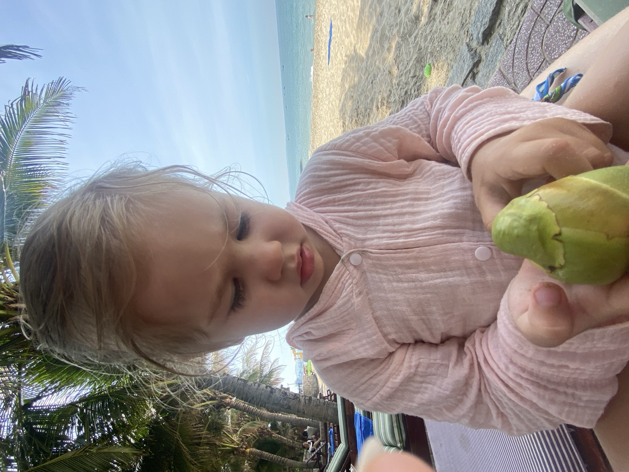A young child with blonde hair is sitting on a striped beach chair, holding a green coconut in their hands. The child is wearing a light pink, long-sleeved garment. The background shows a beach scene with palm trees, lounge chairs, sand, and the ocean under a clear blue sky. In the distance, there are boats on the water and a few people on the beach. The scene is consistent with a location in Phu Thuy Ward, Vietnam, known for its beaches and palm trees.