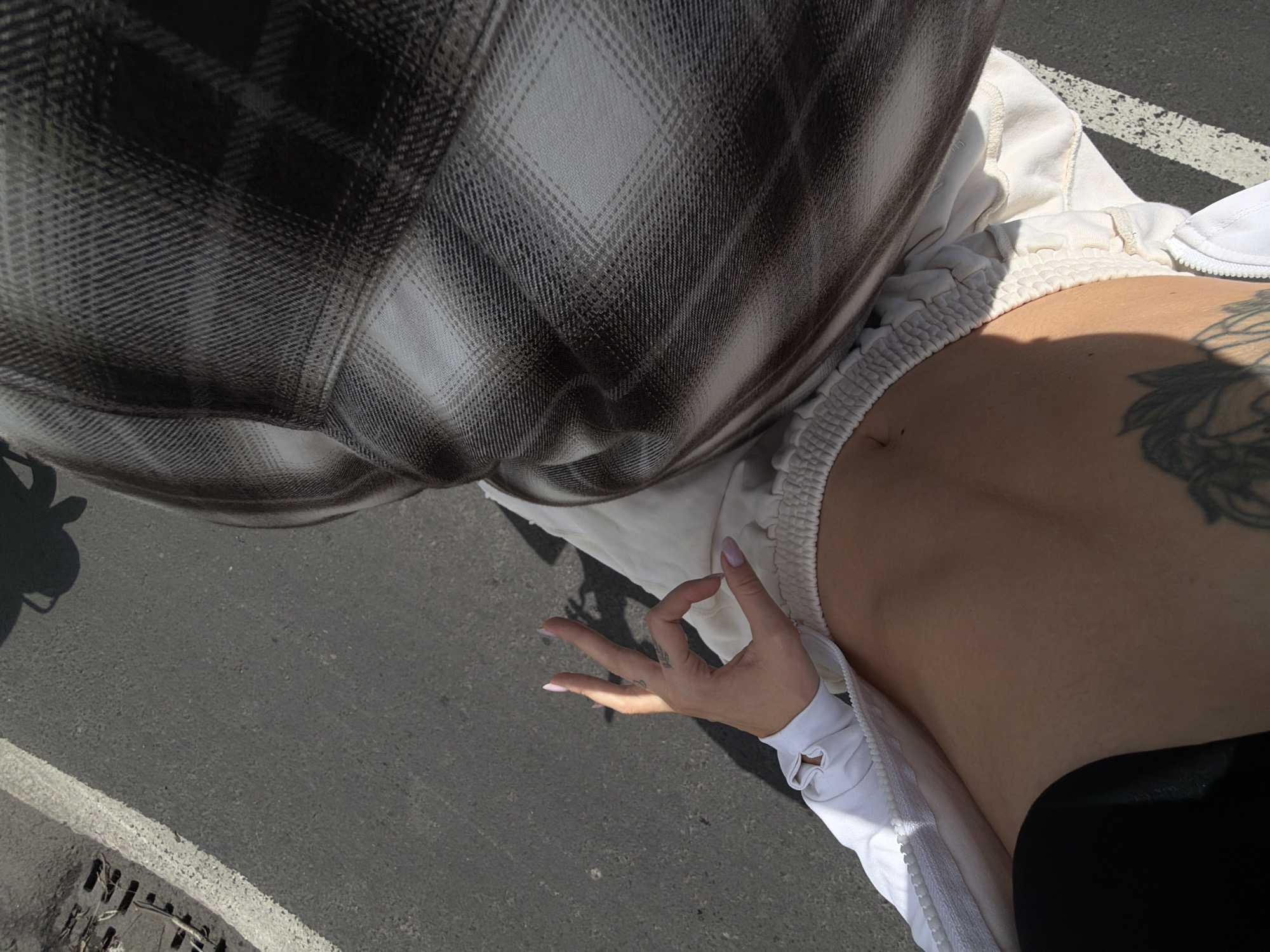 The image is an overhead shot of a person's midsection and hand. The person is wearing a white zip-up jacket over light-colored sweatpants. A shadow of a person wearing a plaid shirt is cast on the ground. The hand is making an "okay" gesture. The background is asphalt with a white line and a drain grate. The scene appears to be outdoors, possibly in a parking lot. The location is specified as North Kuta, Indonesia.