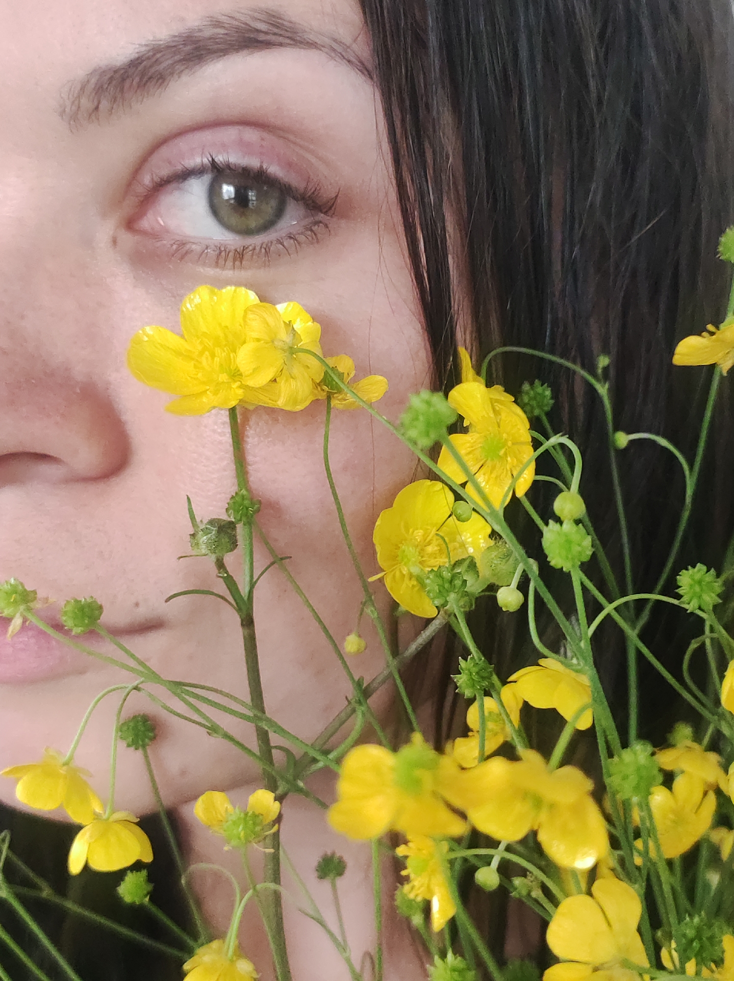 A close-up portrait captures the left profile of an individual's face, partially obscured by a dense cluster of yellow flowers and green foliage. The subject's left eye, situated in the upper-left quadrant of the frame, displays a green iris with a dark pupil, framed by a dark brown eyebrow and visible eyelashes. The skin appears light-toned with visible pores and texture on the cheek and around the eye. The left side of the nose, including the nostril, and a portion of the upper lip are visible in the lower-left area. Dark, straight hair extends vertically along the right edge of the composition. The flowers are small, bright yellow, and possess multiple petals with distinct green centers, resembling buttercups. Numerous spherical green buds and thin green stems are also prominent throughout the floral arrangement. These botanical elements are positioned between the camera and the face, creating varying planes of focus across the image. The individual's gaze is directed towards the viewer. This scene is located in Фоки, Russia.