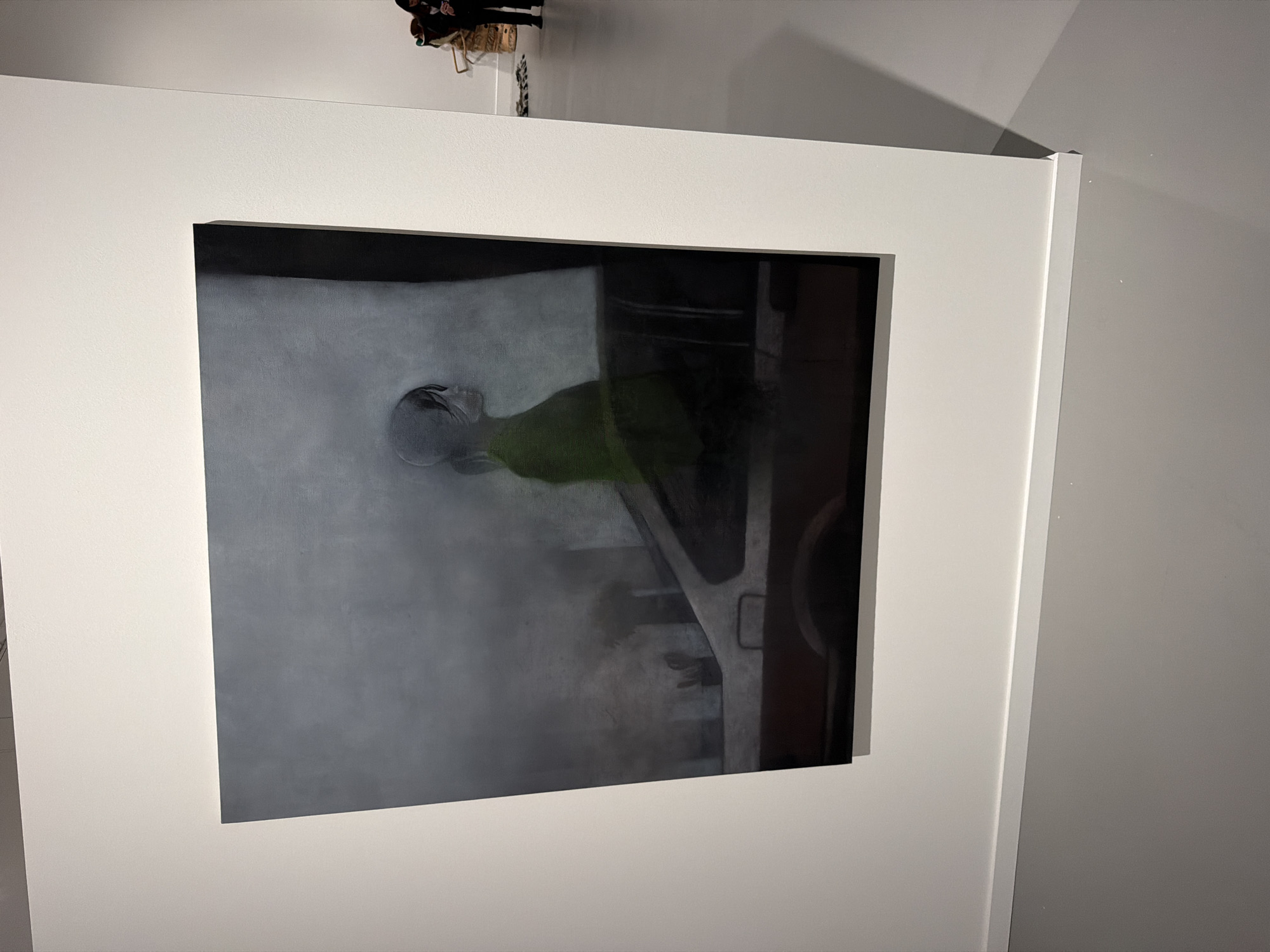 An indoor photograph, rotated counter-clockwise, displays a framed painting mounted on a white wall, likely within an art gallery or exhibition space. The painting features a figure lying horizontally, with its head positioned towards the left side of the canvas. The figure's head and visible limbs are rendered in muted grey tones, while its torso is a distinct, vibrant green. The background of the artwork transitions from a lighter, hazy grey on the left to darker, abstract shapes and structural elements on the right, which could suggest machinery or architectural forms. The painting is encased in a simple, thick white frame. In the upper left corner of the overall image, partially visible, are the heads of two individuals, appearing to observe the artwork. One person has dark hair and wears a patterned garment. The walls are plain white, and the lighting is even, typical of an exhibition environment. No text is visible in the image.