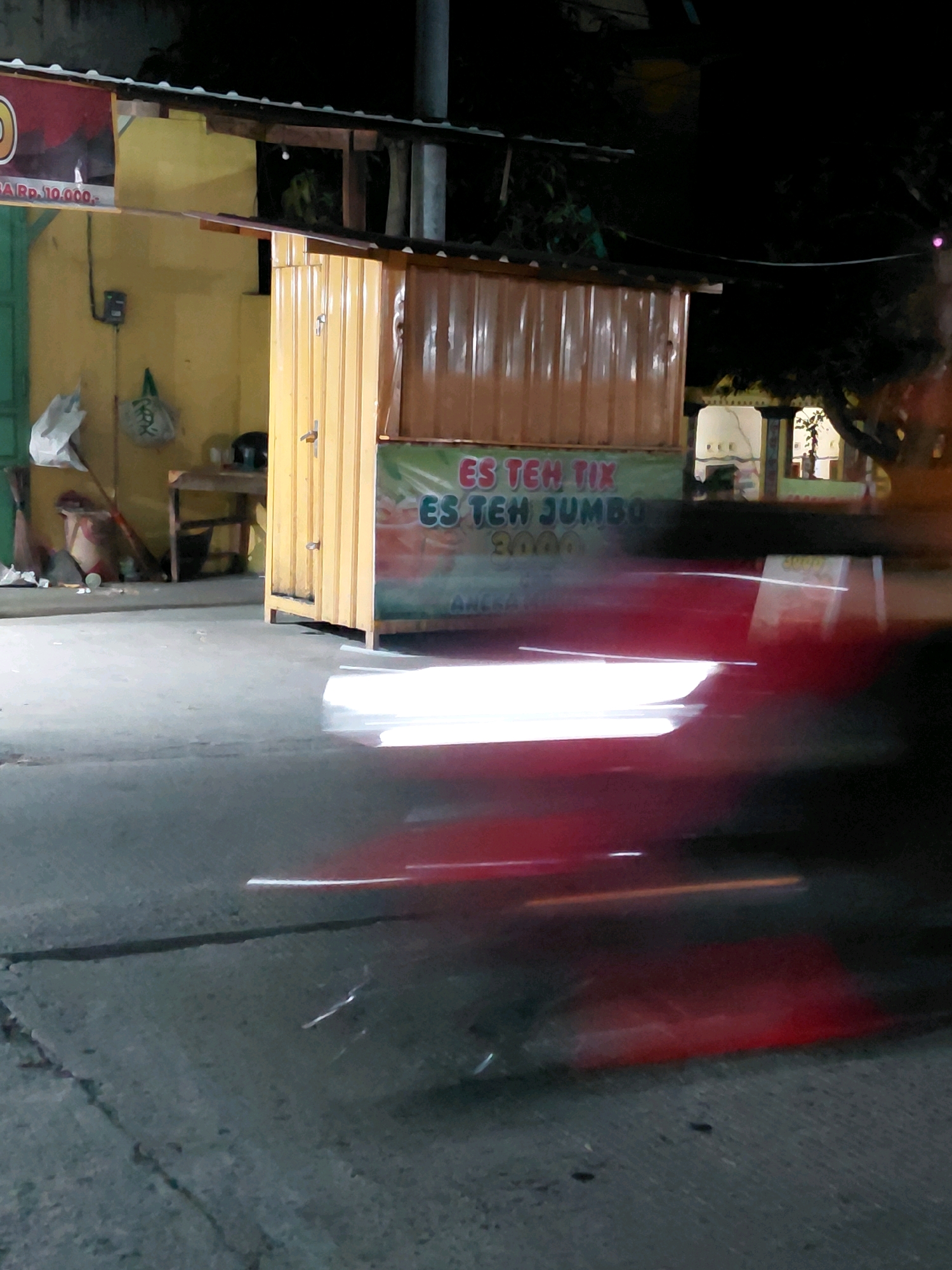 The image shows a roadside scene at night in Babadan, Indonesia, with a food stall and blurred motion. A yellow and brown corrugated metal food stall is situated in the center of the frame. The stall has a sign with text that is partially obscured by motion blur, but "ES TEH TIK" and "JUMBO" are legible. To the left of the stall is a yellow wall with some items hanging, including a white plastic bag and a green bag with writing. Behind the stall, the scene is dark, with some greenery and a dimly lit window visible. The foreground of the image is dominated by a paved surface, likely a road or sidewalk, with a distinct motion blur of red and white streaks indicating a fast-moving vehicle or person. The overall lighting suggests it is nighttime, with artificial lights illuminating the stall.