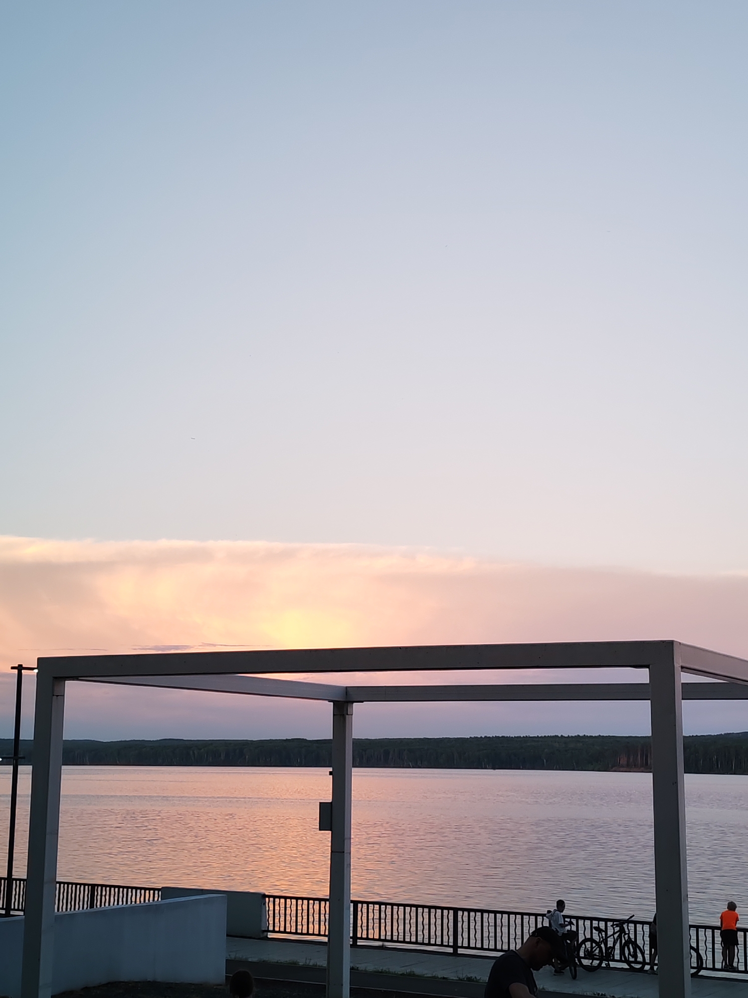 The image depicts a waterfront scene at sunset in Chaikovsky, Russia. A large, calm body of water, likely the Kama River, stretches across the middle ground, reflecting the soft pink and orange hues of the sky and clouds. A dense forest lines the opposite bank. In the foreground, a modern, white, open-air structure with a roof supported by pillars frames the view of the water. A black metal railing runs along the waterfront promenade. Several individuals are visible on the promenade. Two bicycles are parked near the railing, and a person in an orange shirt stands at the far right, looking out at the water. Another person wearing a dark shirt and hat is partially visible in the lower left corner, looking down. The sky above is a gradient of pale blue, transitioning to warmer tones near the horizon where the sun is setting, with a band of soft pink and peach-colored clouds.