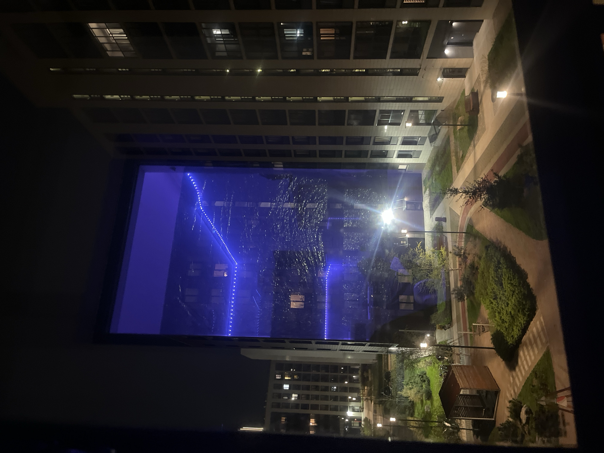 View from an elevated vantage point, possibly a high-rise apartment, at night. Focus on a courtyard surrounded by multi-story buildings in Saint Petersburg, Russia. The courtyard features walkways, vegetation, benches, and a small wooden structure (possibly a gazebo or shelter). The lower portion of the image shows a building with illuminated windows. Across the courtyard is another building, also with illuminated windows. A water fountain or decorative feature is adorned with blue LED lights. Street lamps illuminate the walkways. No people are clearly visible.