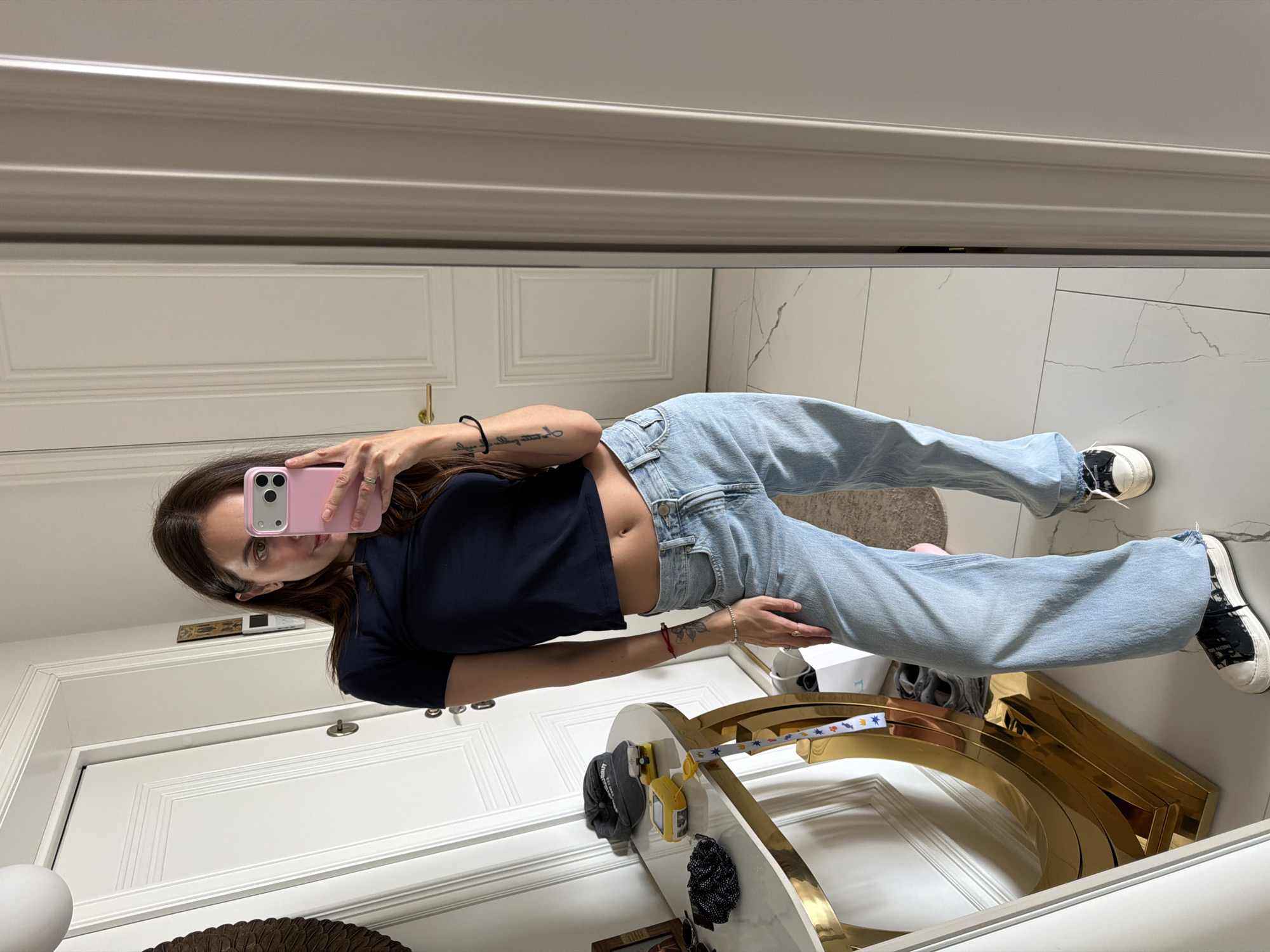 A woman is taking a selfie in a mirror in what appears to be a home interior. She is wearing a black crop top and light blue jeans, with black and white sneakers. The woman is holding a pink iPhone. The background includes a white door, white marble-patterned flooring, and a gold-colored arched decorative piece. The location cannot be determined as the image does not provide enough location information to identify that the location is in Krasnoyarsk, Russia.