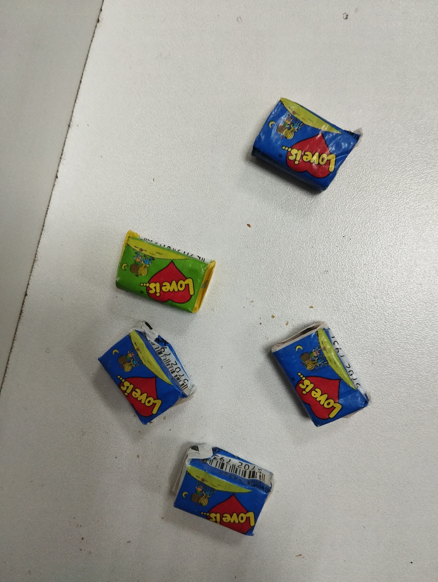The image shows five pieces of "Love is..." gum wrappers scattered on a white surface. Four wrappers are blue with a red heart and the "Love is..." logo, with one wrapper showing the barcode "5702 1931". The remaining wrapper is green with a red heart and the "Love is..." logo.