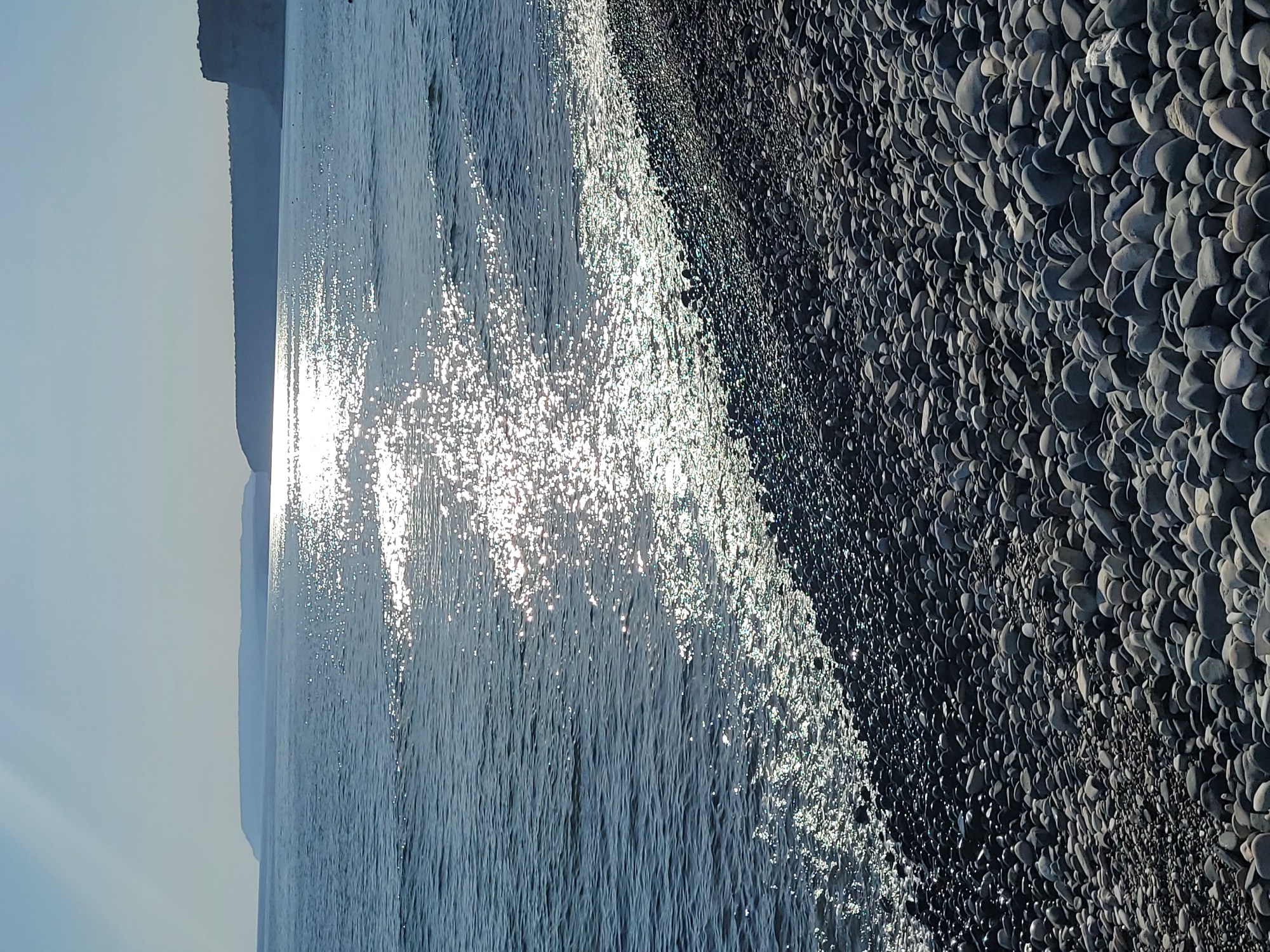 The image depicts a sunny day at a pebble beach in Episkopi, Cyprus. The foreground is dominated by a dense scattering of smooth, grey and darker-toned pebbles, filling the lower half of the frame. The sea occupies the middle ground, its surface shimmering with reflections of the bright sun. Gentle waves lap at the shore, creating a line of sparkling foam where they meet the pebbles. In the background, a hazy, low-lying coastline stretches across the horizon, its form softened by the distance and atmospheric conditions. The sky above is a clear, pale blue, with a faint sunbeam visible on the upper left. No people are visible in the scene.