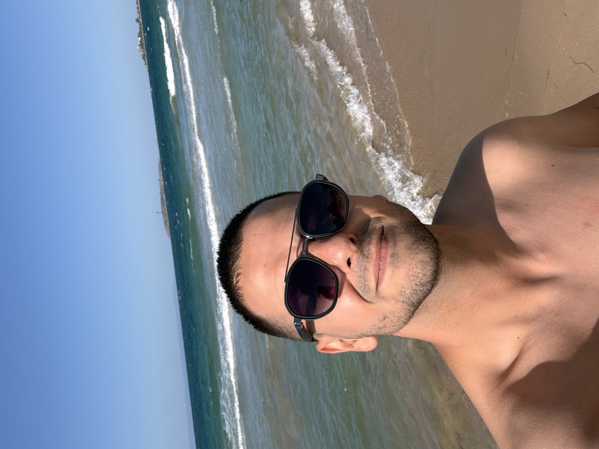 A man is taking a selfie on a sandy beach. The man is wearing sunglasses and is shirtless. The ocean is visible in the background with waves gently rolling onto the shore. The sky is clear and blue. The location appears to be a coastal area, likely a popular tourist destination.