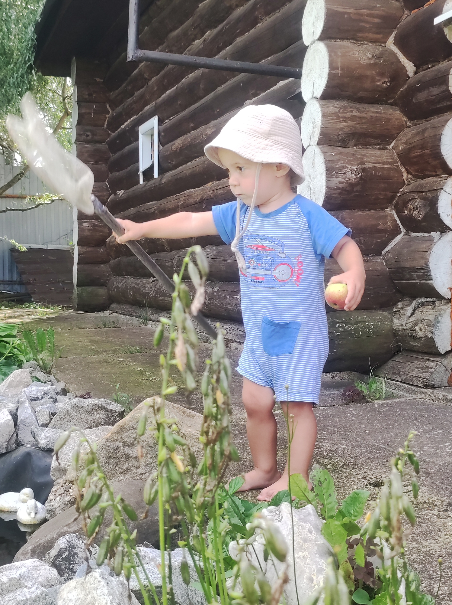 A young child wearing a bucket hat and a blue and white striped romper stands on a concrete surface in front of a log cabin. The child is holding a butterfly net in their right hand and an apple in their left hand. In the foreground, there are rocks and some greenery. To the left of the child, there is a small pond with two white decorative ducks. The background features the textured wall of the log cabin.