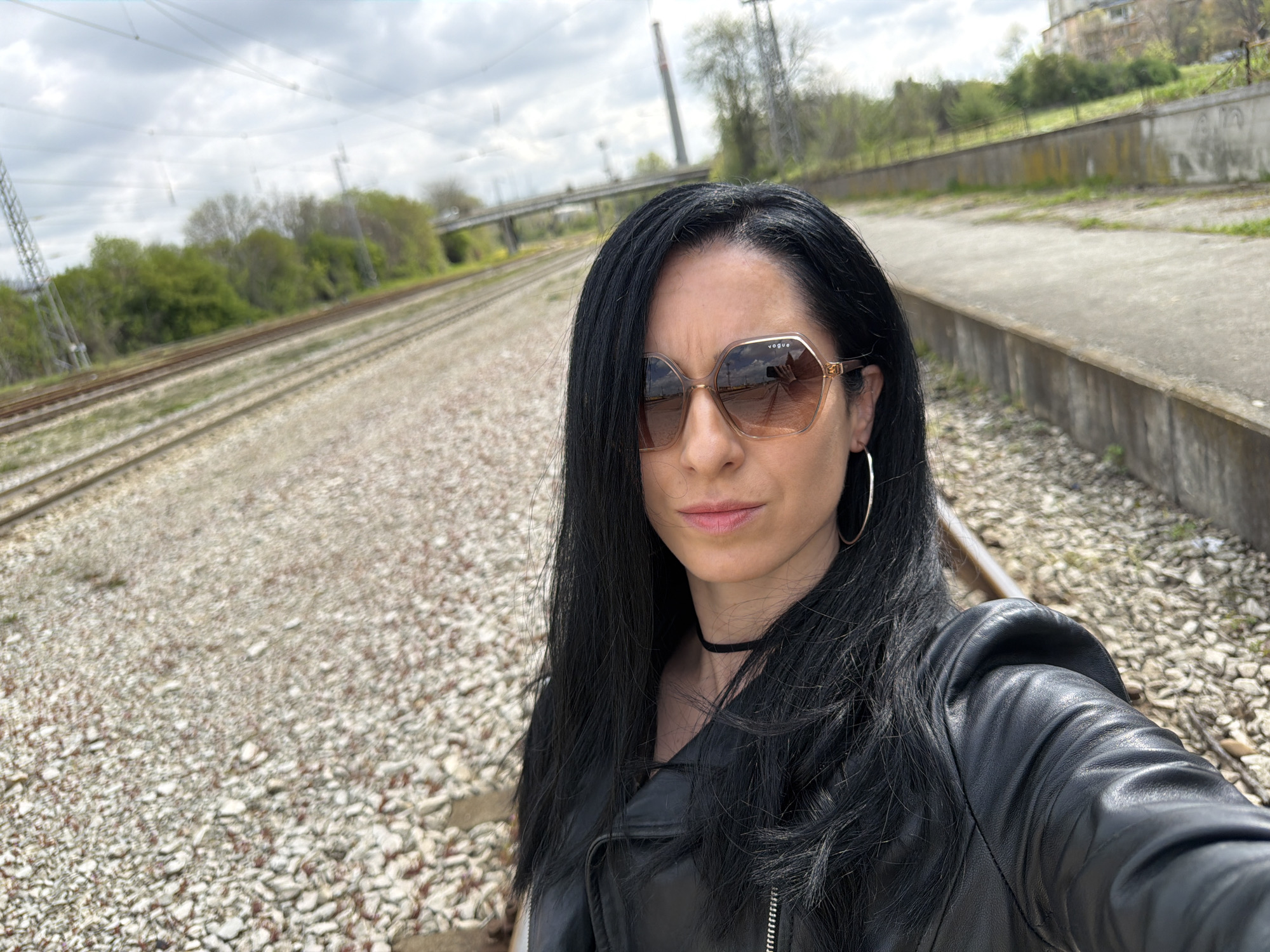 The image shows a woman standing on a gravel area adjacent to railroad tracks. She is wearing sunglasses, a black leather jacket, and a choker. The tracks extend into the distance, with a bridge visible in the background. The location is Pleven, Bulgaria, identifiable from the distinctive architecture of buildings, trees, and infrastructure elements visible in the background. A concrete barrier runs along the edge of the gravel area, and vegetation is present on the other side of the tracks. The sky is overcast.