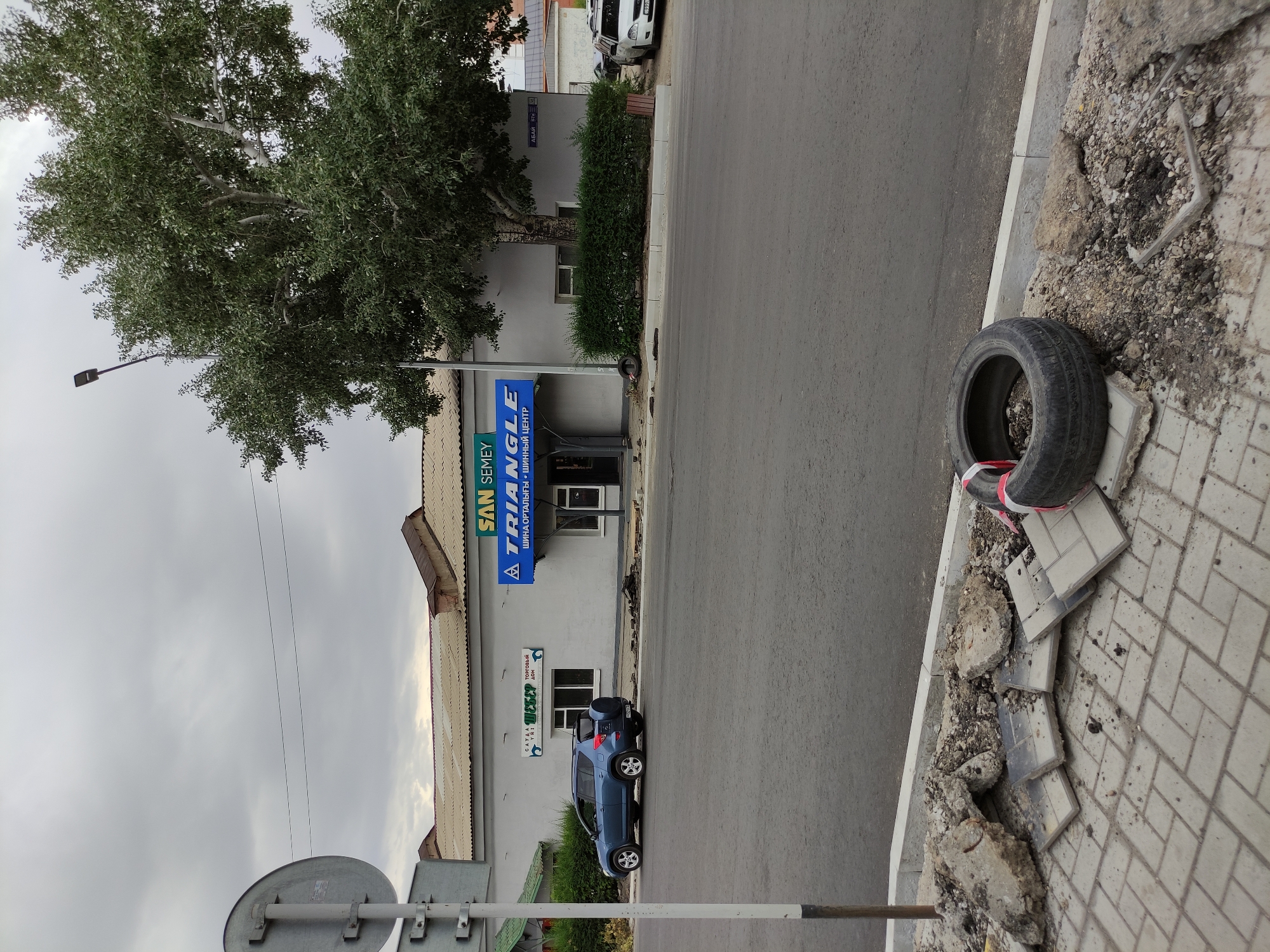 The image shows a street view in Semey, Kazakhstan, under an overcast sky. In the foreground, a sidewalk with broken paving stones and debris is visible. A discarded tire with red and white tape lies on the ground. A gray road runs parallel to the sidewalk. Across the road, a building with a light gray facade has two businesses. One is branded "SAN SEMEY," and the other has a blue sign that reads "TRIANGLE" in English and Cyrillic, indicating a tire center. A blue SUV is parked in front of the building, and a white car is parked further down the street. A street lamp is positioned next to a large tree with lush green foliage. A street sign with "АБАЙ 97a" in Cyrillic is mounted on the building.