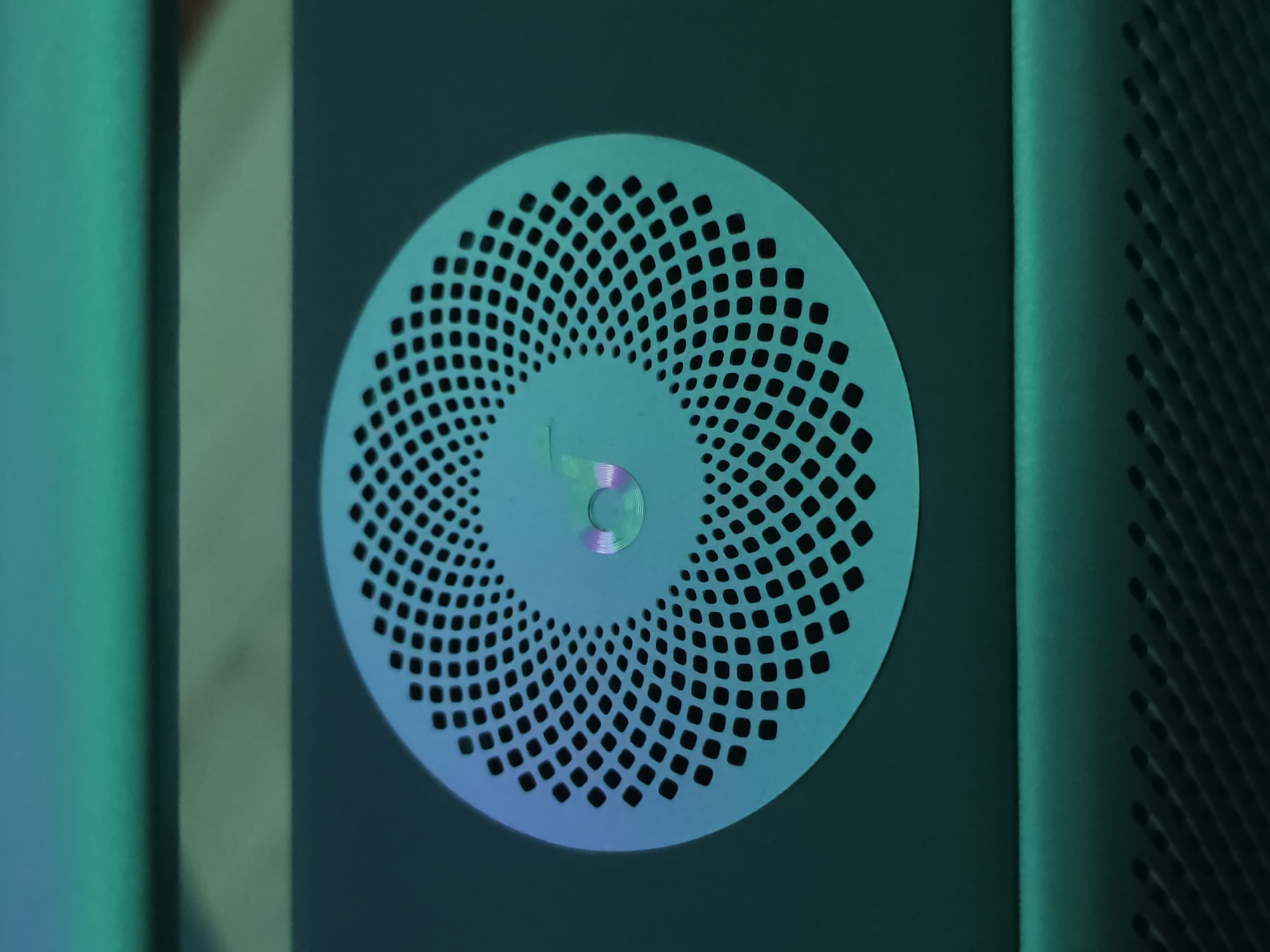 This media file is a close-up, medium shot of a circular vent with a pattern of small squares. The vent is a light teal color and has a circular indentation in the center. The overall tone of the image is dark and moody, with green and blue lighting illuminating the vent from the side. To the right of the vent, there is another textured surface with a similar pattern of squares, suggesting it's part of a larger object or structure. The left side of the image shows a smooth, dark surface with a vertical line of bright green light. The lighting creates a futuristic or industrial aesthetic. There is no discernible text or human presence in the image. The scene appears to be indoors, possibly a studio or a controlled environment, given the artificial lighting and focus on the object.