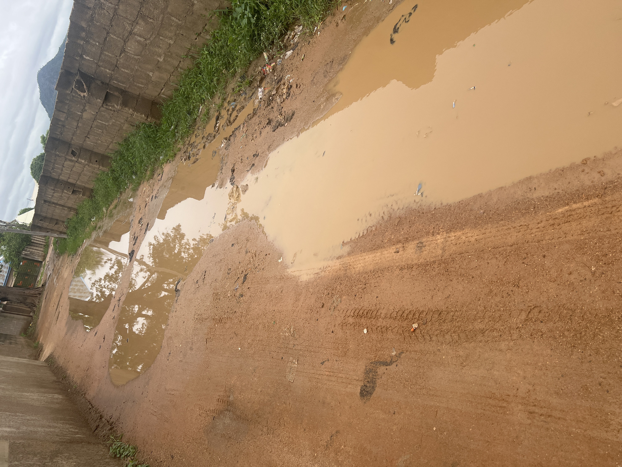 A dirt road in Jalingo, Nigeria, is partially flooded with muddy water, forming large puddles that reflect the sky and surrounding trees. The road is flanked by a concrete wall on the right and a building wall on the left. There are tire tracks visible on the muddy ground. Some litter is scattered along the edge of the road next to the wall. In the background, a hill is visible under a cloudy sky.