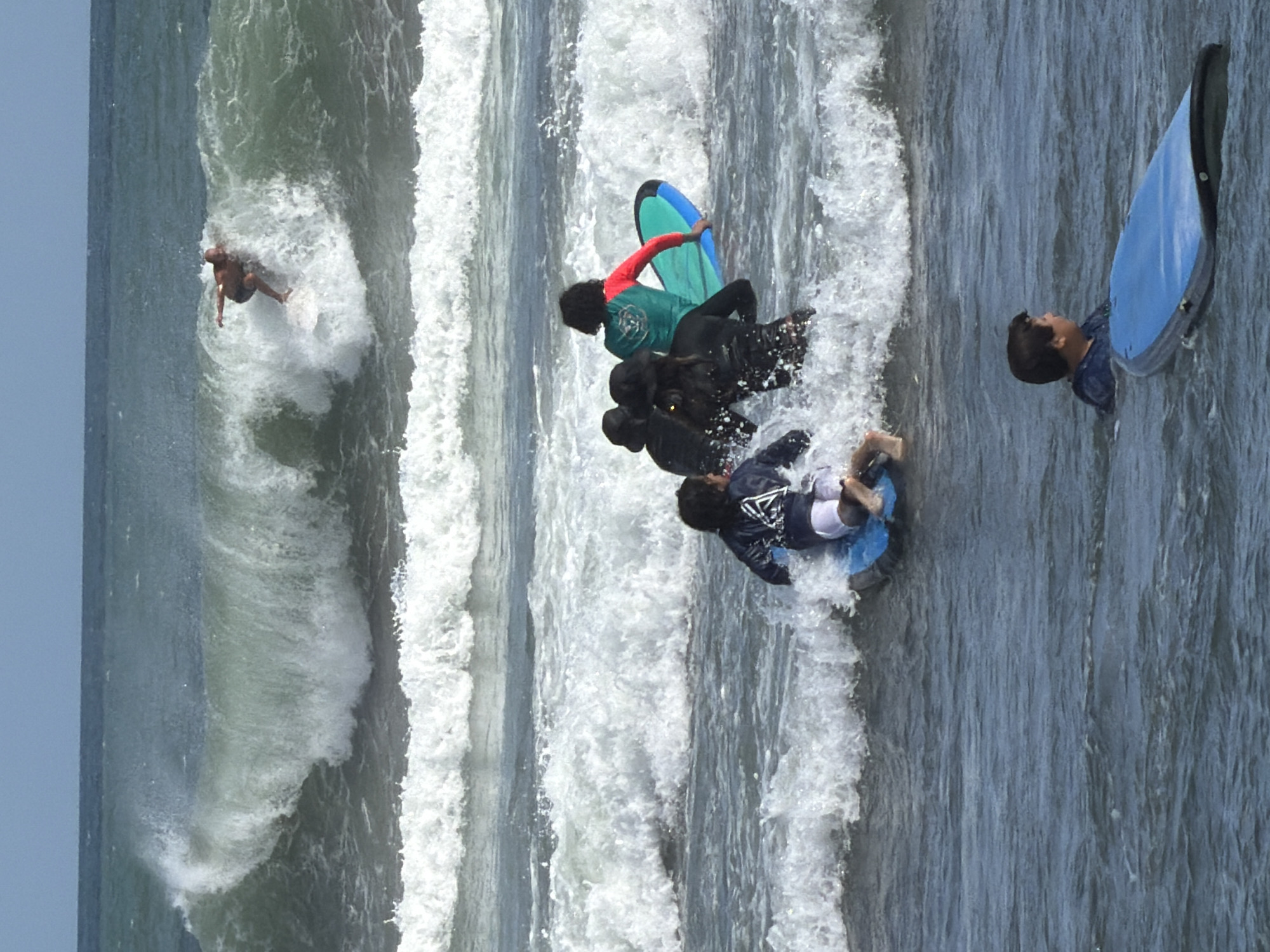 The image depicts a group of surfers in the ocean near North Kuta, Indonesia. In the background, a male surfer is riding a wave. In the mid-ground, several individuals, some appearing to be children, are on surfboards in choppy water, with one child in the foreground on a blue surfboard. The environment is characterized by waves breaking and white foam. The sky is overcast.