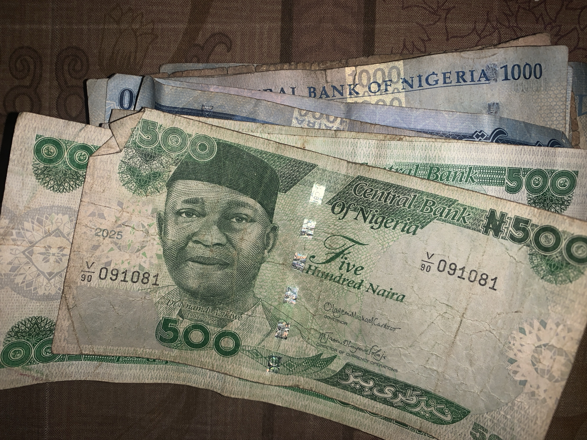 A close-up, overhead shot displays several used Nigerian Naira banknotes arranged on a dark brown, textured fabric surface with a subtle patterned design. The prominent notes are 500 Naira denominations, characterized by their greenish-grey color and visible creases and wear. A 1000 Naira note, with a lighter blueish hue, is partially visible underneath the stack.
The 500 Naira notes feature a portrait of Dr. Nnamdi Azikiwe. Visible security features include small reflective holograms. The banknotes appear to be in circulation, showing signs of frequent handling.
Visible text on the notes includes "Central Bank of Nigeria," "N500," and "Five Hundred Naira." The name "DR Nnamdi Azikiwe" with the dates "1904-1996" is legible below his portrait. Signatures of "Olayemi Michael Cardoso GOVERNOR" and "Jamii Olayemi Solaja DIRECTOR OF CURRENCY OPERATIONS" are also present. Serial numbers like "V 091081" appear multiple times, alongside the number "2025" on one note. The 1000 Naira note clearly shows "1000" and "BANK OF NIGERIA." Some Arabic script is also visible on the 500 Naira note. The scene suggests an indoor setting in Rafin Par, Nigeria.