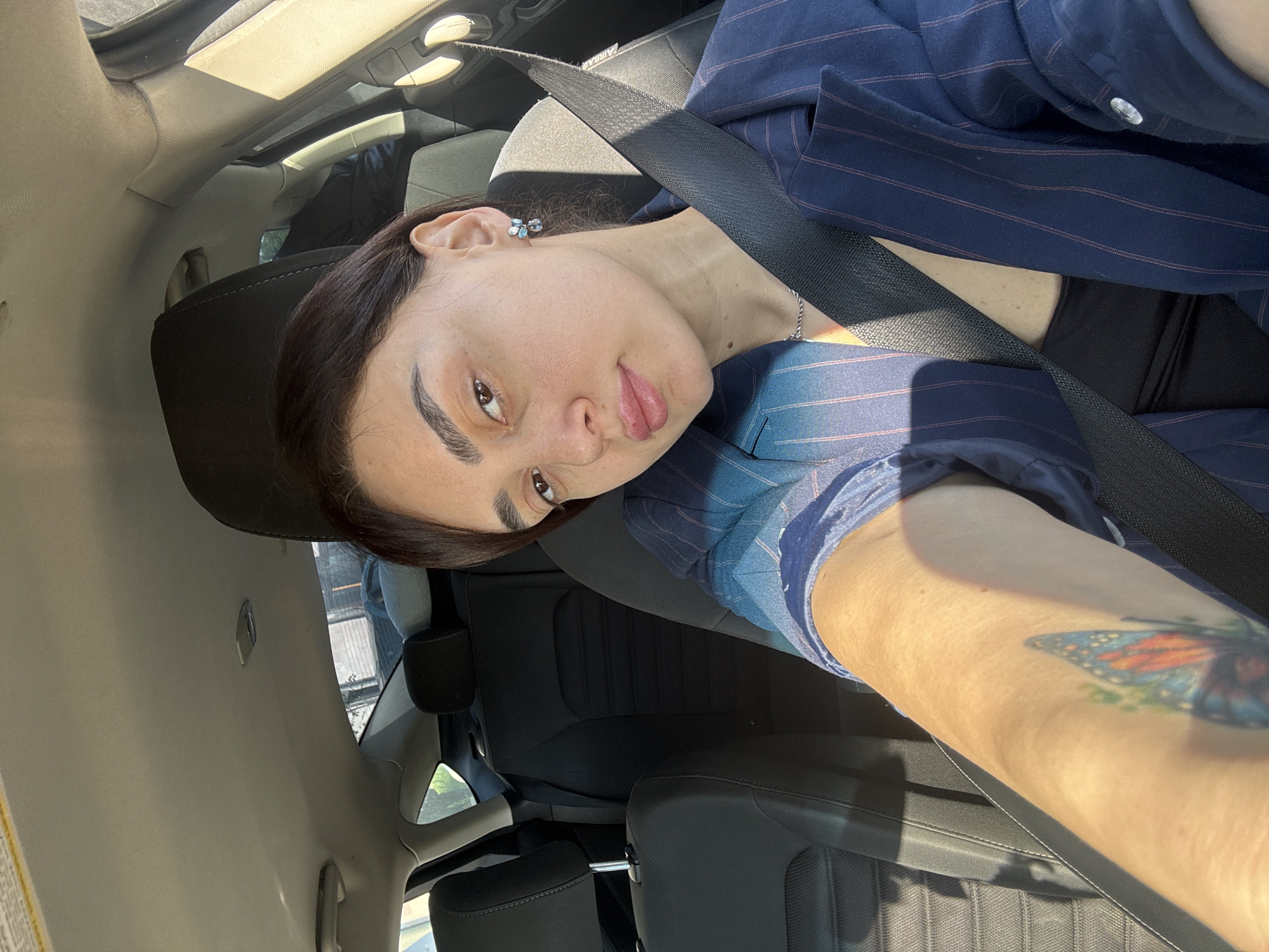 This portrait-style image, oriented with the subject's head to the right, captures a young woman seated in what appears to be the front passenger seat of a car. She has dark hair, distinct dark eyebrows, and brown eyes, looking directly at the camera with a neutral to slightly pouty expression. She wears a dark blue blazer with faint pinstripes over a dark-colored top. A small, light-colored earring, possibly featuring blue or silver stones in a floral or star-like shape, is visible in her right ear, along with a thin silver-colored chain necklace. Across her chest, a black seatbelt is fastened. On her right arm, a vibrant tattoo depicting a monarch butterfly wing, featuring orange, blue, and green hues, is clearly visible.
The interior of the car features dark-colored seats and a light-colored headliner. Bright daylight illuminates the scene, casting strong shadows within the cabin, indicating a sunny day. A sunroof or panoramic roof is partially visible, allowing light to stream in. Blurred elements seen through the car windows suggest an outdoor background. The word "AIRBAG" is faintly discernible on the car's interior panel above the seatbelt. The image context places the location in Tbilisi, Georgia.