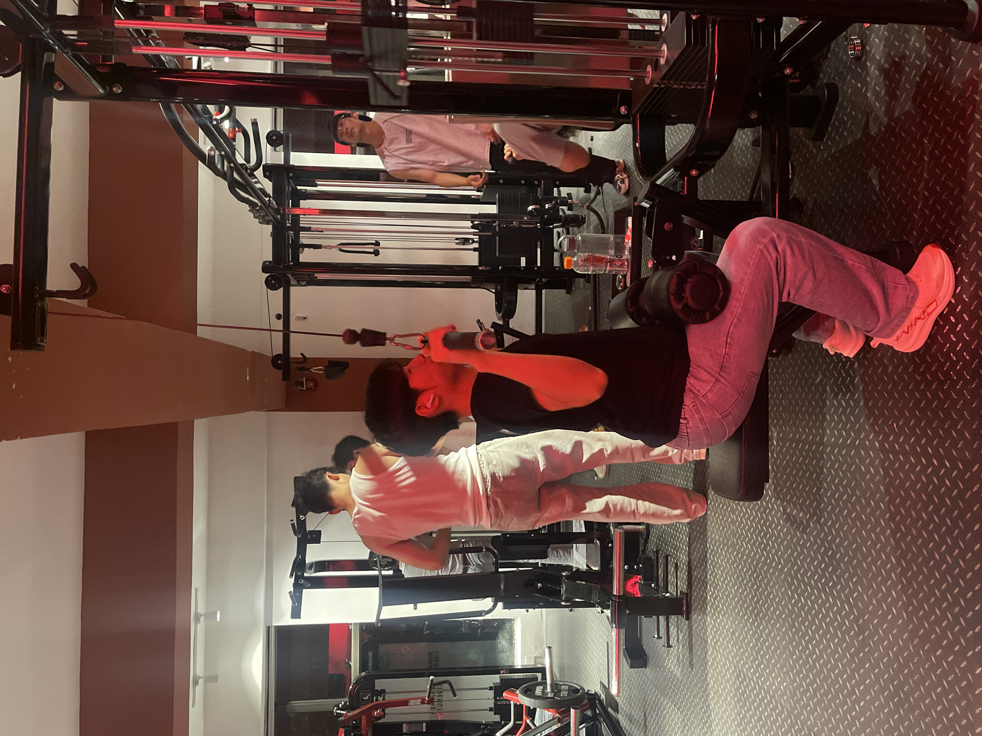 Location: Gym in Chongqing High-tech Zone, China.
Scene: Several individuals are engaged in physical exercise using various weight machines. One person is seated, performing leg extensions. Another person is supine, possibly using a machine for chest or shoulder exercises. A third person is visible in the background, potentially using a pull-up machine. The gym has a dark aesthetic with black equipment and a patterned floor. Red lighting accents the room. Water bottles are placed near the equipment.