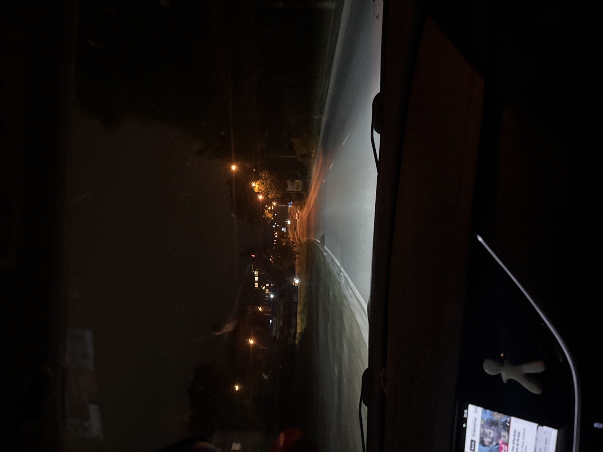 The image depicts a nighttime scene from the interior of a vehicle, looking forward through the windshield onto a dimly lit road in Varna, Bulgaria. The time, visible on an infotainment screen, is 22:45.
The asphalt road, illuminated by the vehicle's headlights, shows white lane markings and curves gently to the right. On the left side of the road, a grassy bank slopes upwards towards several buildings, some of which display illuminated windows. Multiple streetlights with warm, orange illumination line this side of the road. On the right, dense dark foliage borders the road, interspersed with additional streetlights further in the distance.
The vehicle's dark dashboard occupies the lower portion of the frame. On the lower-left, a digital infotainment screen displays text and images, with the time "22:45" prominently visible. Adjacent to the screen, a small, light-colored, stylized human figurine is positioned. A thin white cable is visible traversing the dashboard from the lower-left towards the center-right. Reflections, possibly from the vehicle's interior or a passenger, are faintly discernible in the upper left quadrant of the windshield. The overall environment is dark, with artificial light sources providing primary illumination. No specific individuals or explicit actions are clearly visible beyond the implied presence of an occupant taking the photograph.