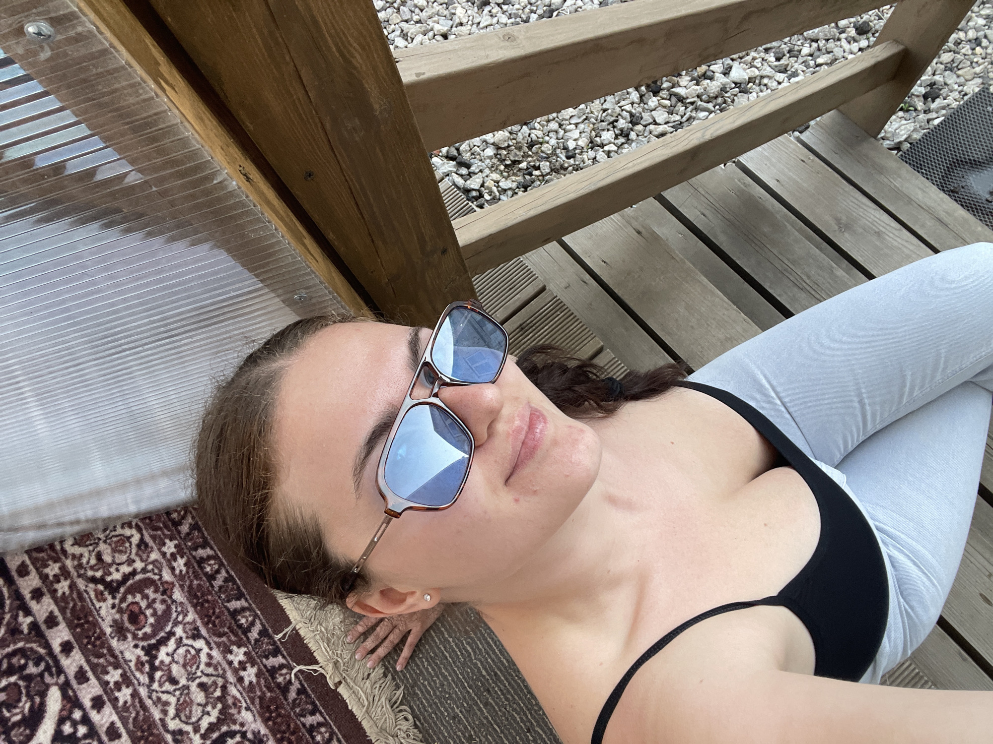 A woman is lying on a wooden platform in Мосметаллоконструкция Онт, Russia, wearing sunglasses, a black top, and light blue jeans. She is lying on a patterned rug, with a wooden structure above her and gravel visible through the gaps in the wooden platform.