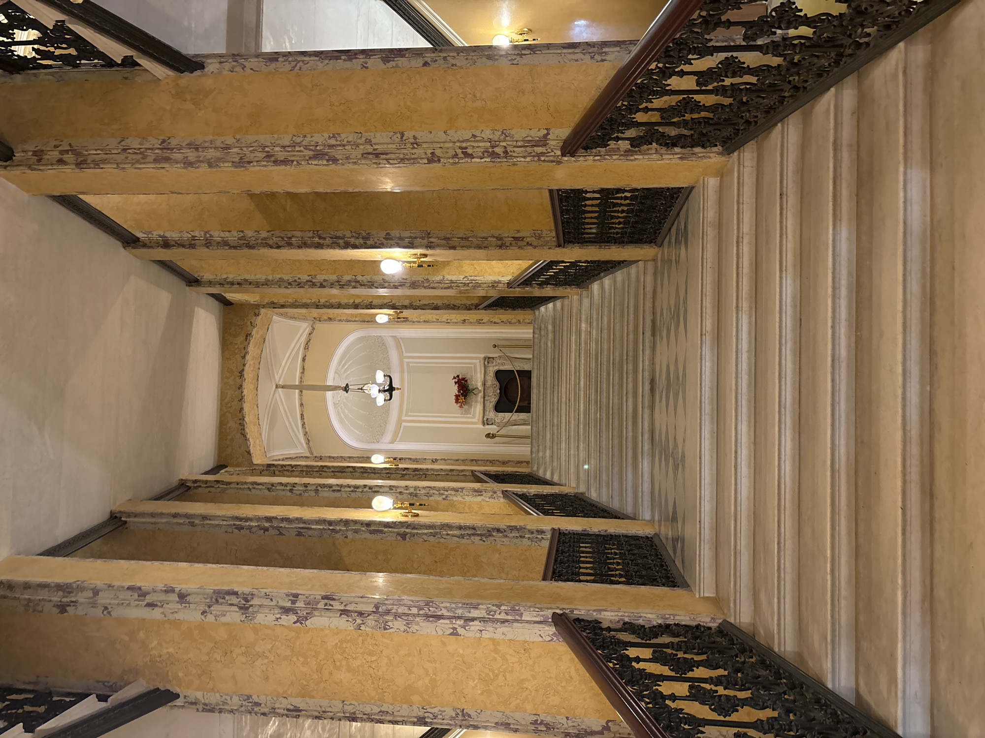 This is a frontal, eye-level, interior shot of a grand marble staircase in Gatchina, Russia, likely within a historic building such as the Gatchina Palace. The staircase, with its white marble steps and black wrought-iron railings, ascends towards an arched alcove. The walls are adorned with large, yellow marble columns featuring purple veining, interspersed with decorative sconces holding glowing light bulbs. A checkered floor pattern is visible at the landing before the alcove. At the top of the staircase, an ornate chandelier hangs from a vaulted ceiling, and a fireplace with a decorative urn of red flowers sits in the alcove. The scene is brightly lit, creating an atmosphere of opulence and grandeur.