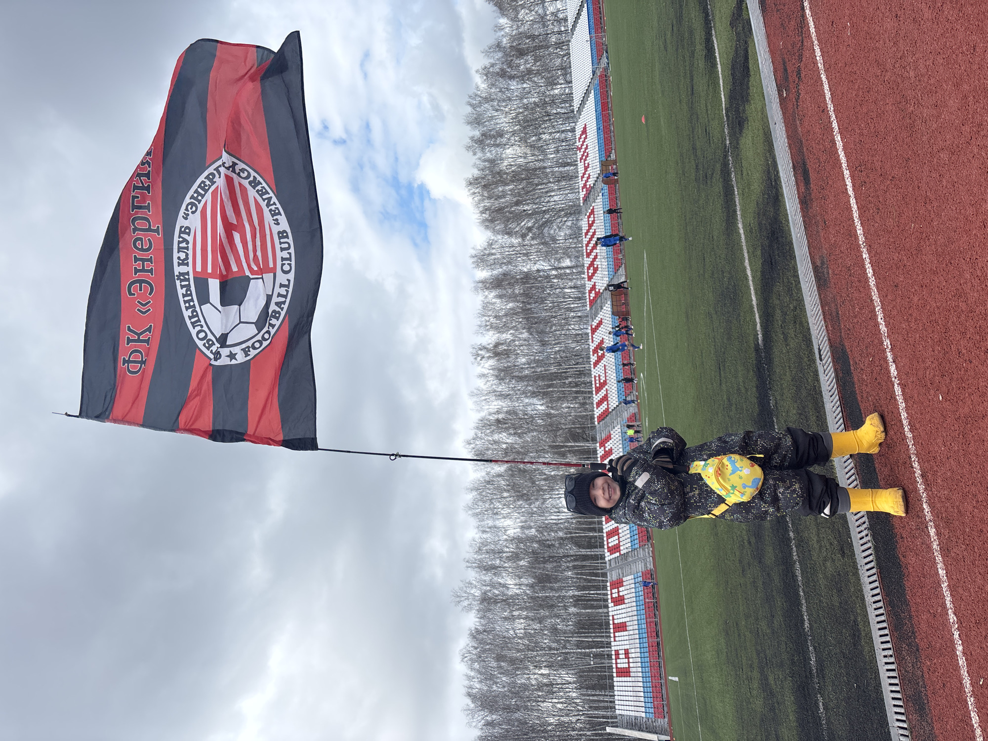 A child in yellow boots stands on a red running track holding a flag of the FC Energiya Chaikovsky football club. The child is wearing a dark patterned snowsuit, black mittens, and a dark hat. The flag has a black and red striped pattern with a circular logo featuring a football and the club name in Russian and English. In the background is a football field, empty stadium seating in Russian national colors, and a line of bare trees against a cloudy sky. The location is likely in Chaikovsky, Russia, based on the football club name.