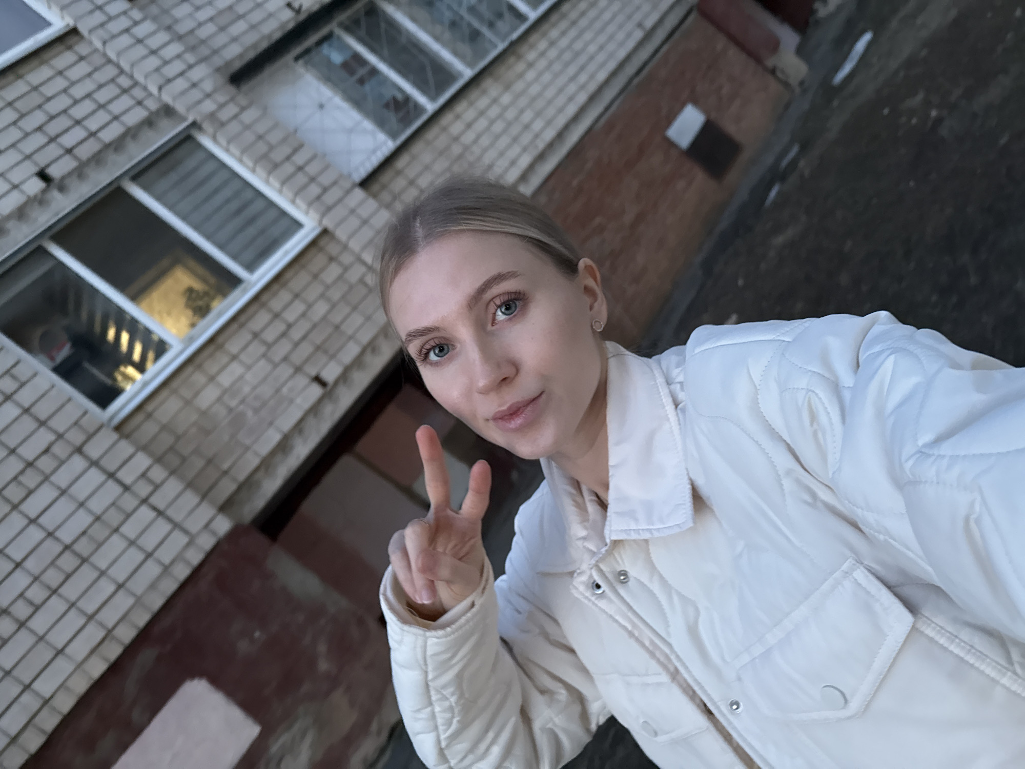 A woman takes a selfie outdoors, holding up a "peace" sign. She has fair skin, blonde hair pulled back, and is wearing a white quilted jacket. The background features a tiled building exterior with windows and a brick portion. The location is Izhevsk, Russia.