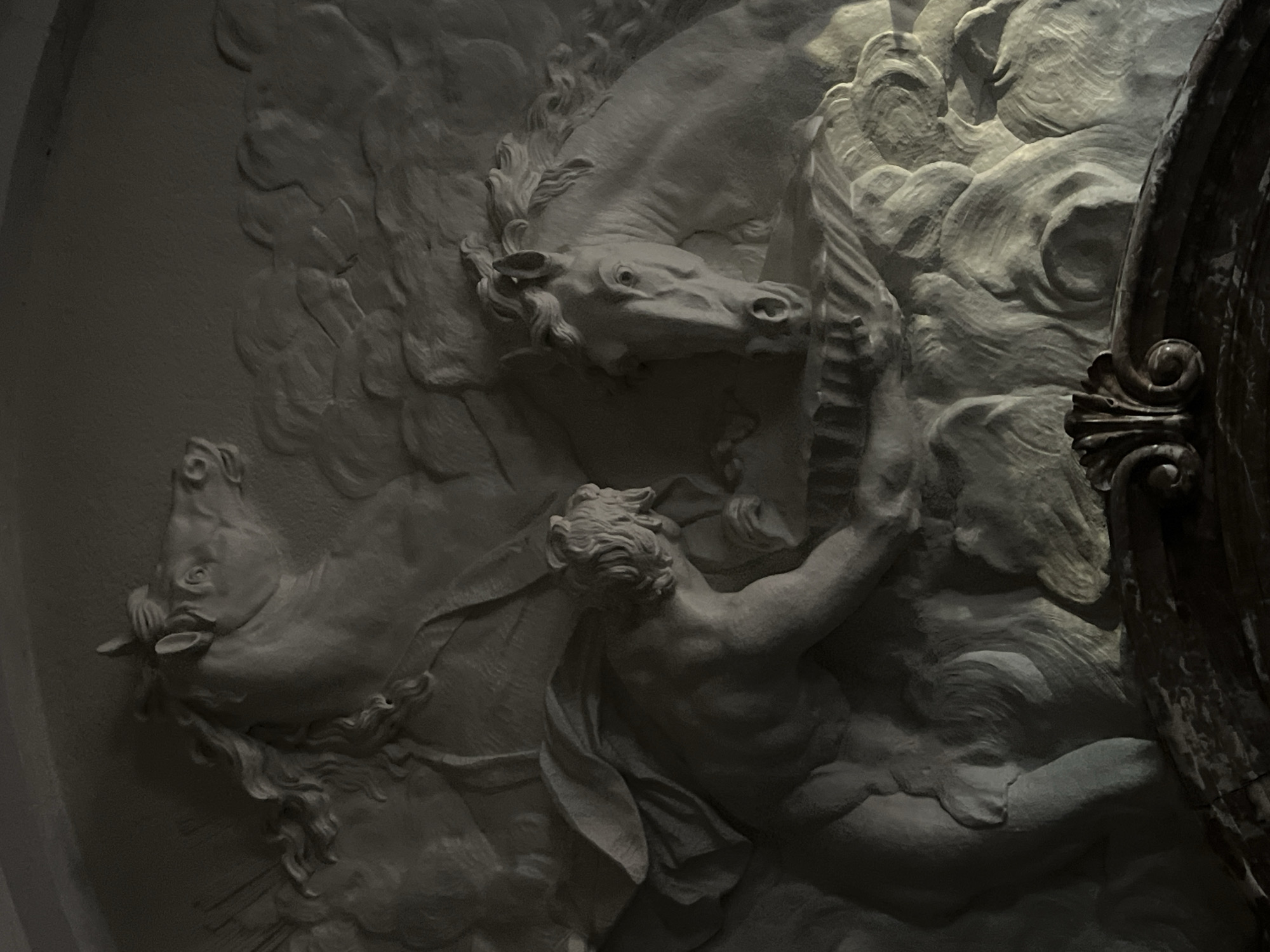 The image shows a section of bas-relief sculpture located in Saint Petersburg, Russia. The relief depicts a classical scene involving figures and horses. A muscular male figure is prominently featured, seemingly in a dynamic pose, with his arm raised. Two horses are also present, their forms conveying a sense of movement and energy. The style is reminiscent of Baroque or Neoclassical art. The texture is stone and it appears the sculpture is on a wall.