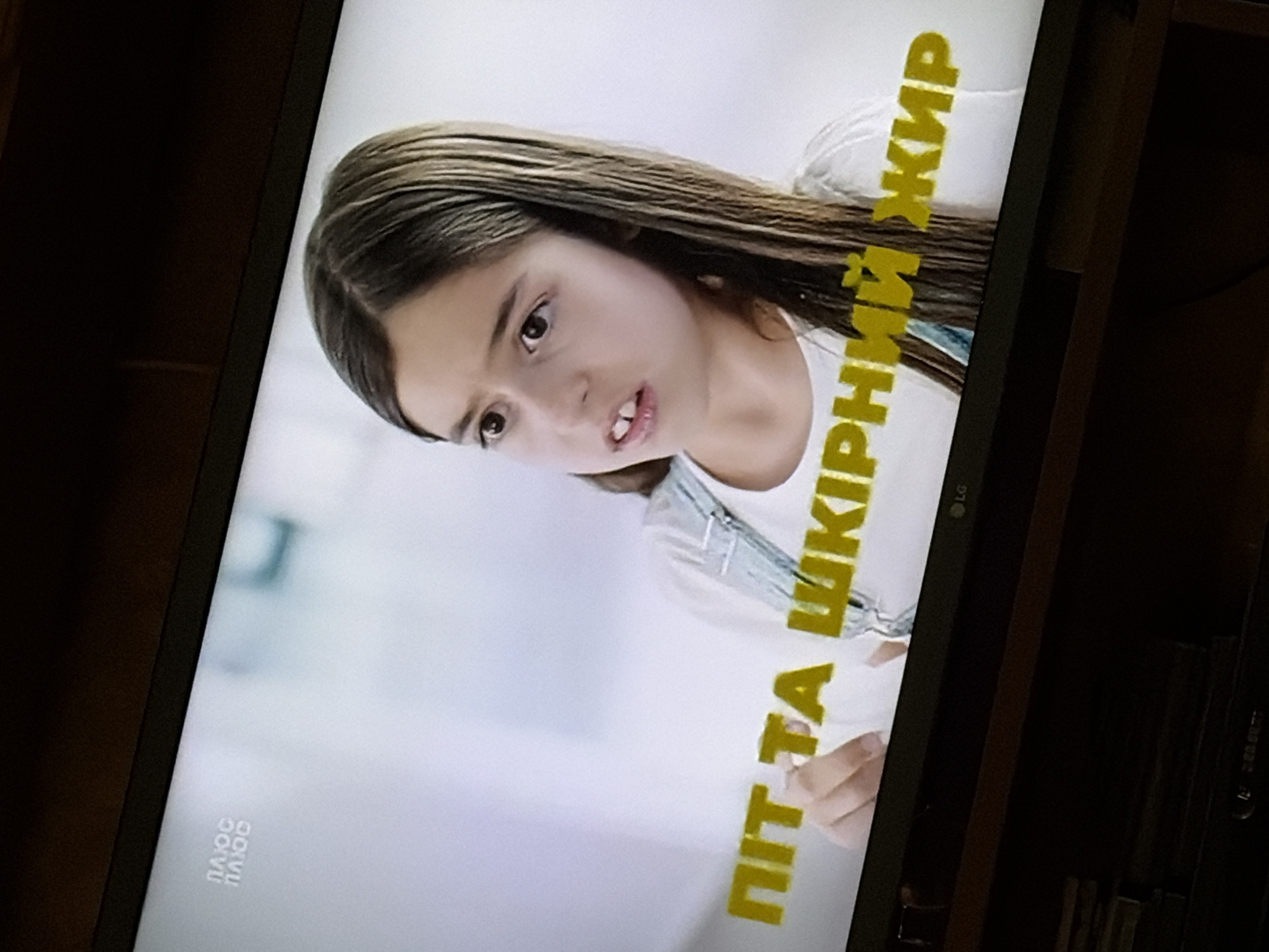 This media file is a photograph taken of a television screen, likely during playback of a video or movie. The primary subject is a young girl with long, dark hair and expressive eyes. She is looking downwards and slightly to her left, with a neutral or slightly curious expression. Her mouth is slightly open, revealing her teeth. She is wearing a white t-shirt and blue overalls.
The background of the image on the screen is a plain, light color, likely white or a very pale gray. This suggests the setting is a studio or a clean, minimalist environment, or it could be a segment of a show focusing on a close-up of the person.
Visible text is present on the screen. In the lower left corner, the text "ПЛЮС ПЛЮС" is displayed in a light color against the light background. On the right side of the screen, running vertically and diagonally upwards, is prominent text in a bright yellow color with a bold, blocky font. This text reads "ПІТ ТА ШКІРНИЙ ЖИР". This text appears to be a title, subtitle, or a key phrase from the content being displayed.
The photograph is taken at an angle, causing the image on the screen to be rotated. The overall lighting is somewhat dim, as is typical when photographing a screen in a darker room. There are reflections on the screen, and the edges of the television set are visible, indicating it is a physical object. The LG logo is visible on the bezel of the TV.
